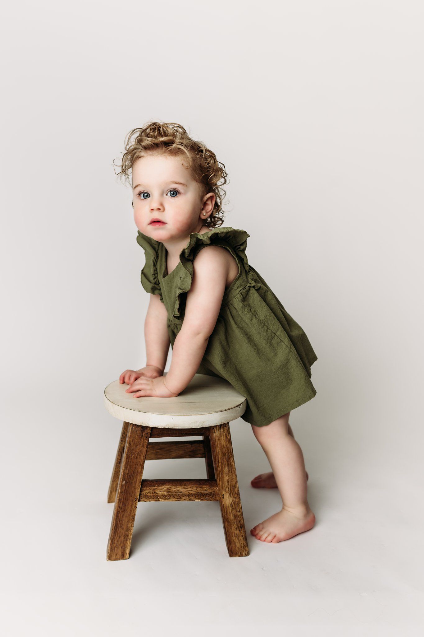 Child in green dress leans on wooden stool against white backdrop.