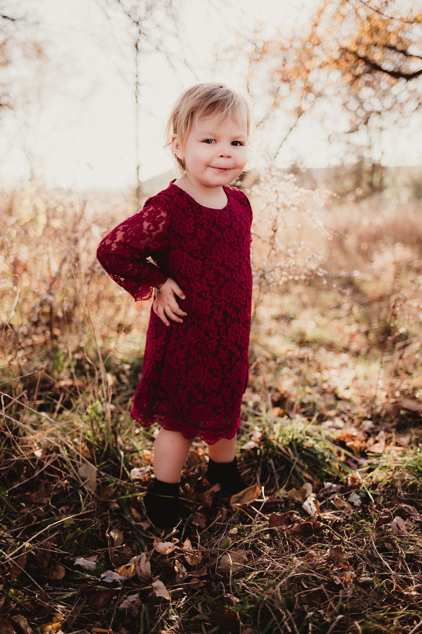 Small child in burgundy lace dress poses confidently among autumn leaves.