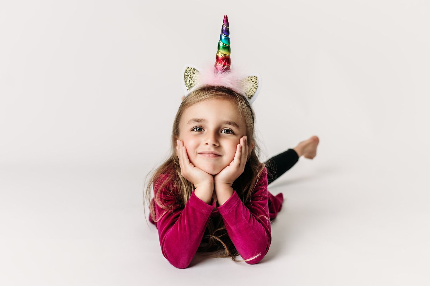 Child wearing unicorn headband and burgundy dress lies on floor with hands under chin.