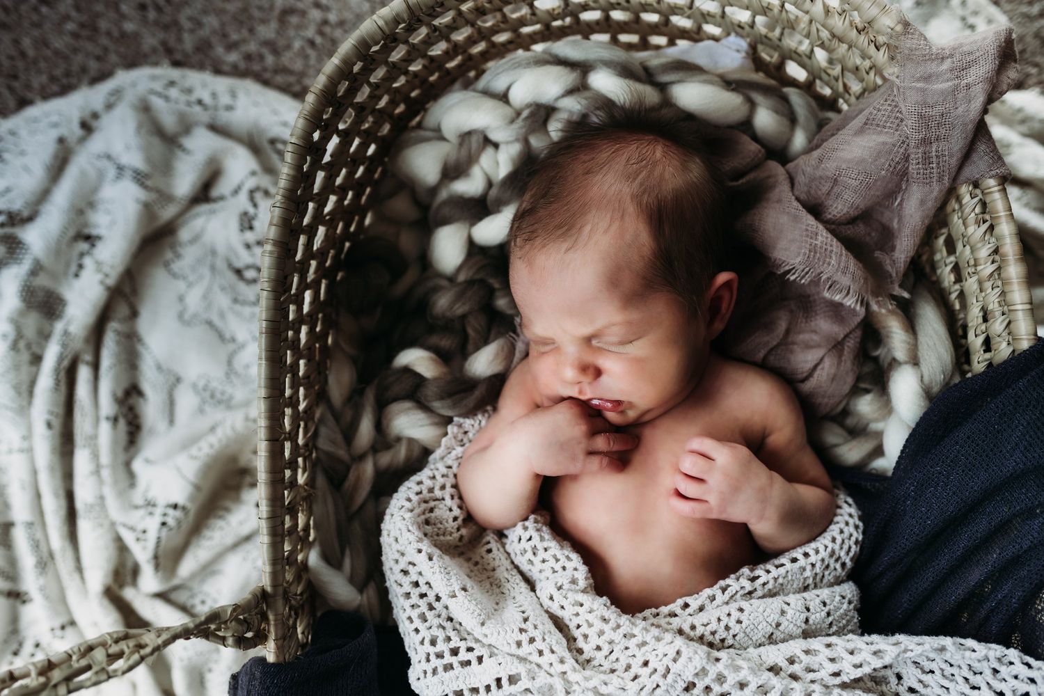 Newborn baby sleeping peacefully in a woven bassinet wrapped in a knit blanket.