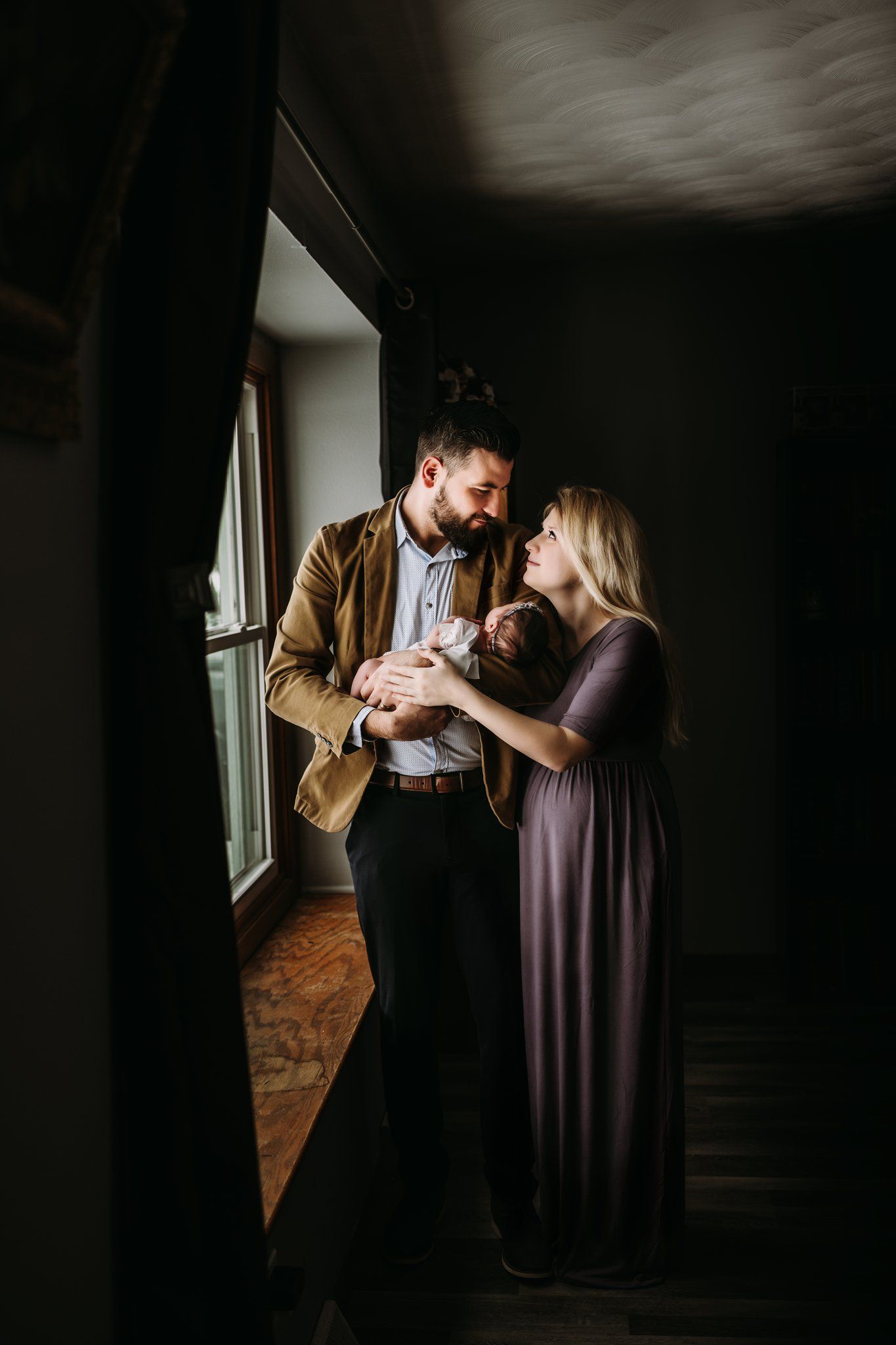 A couple stands in dramatic window light while holding their newborn baby.