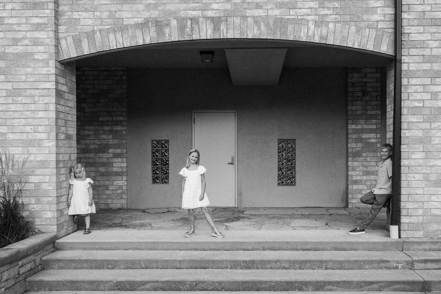 Children in white dresses pose playfully on a stone staircase with brick walls in the background.