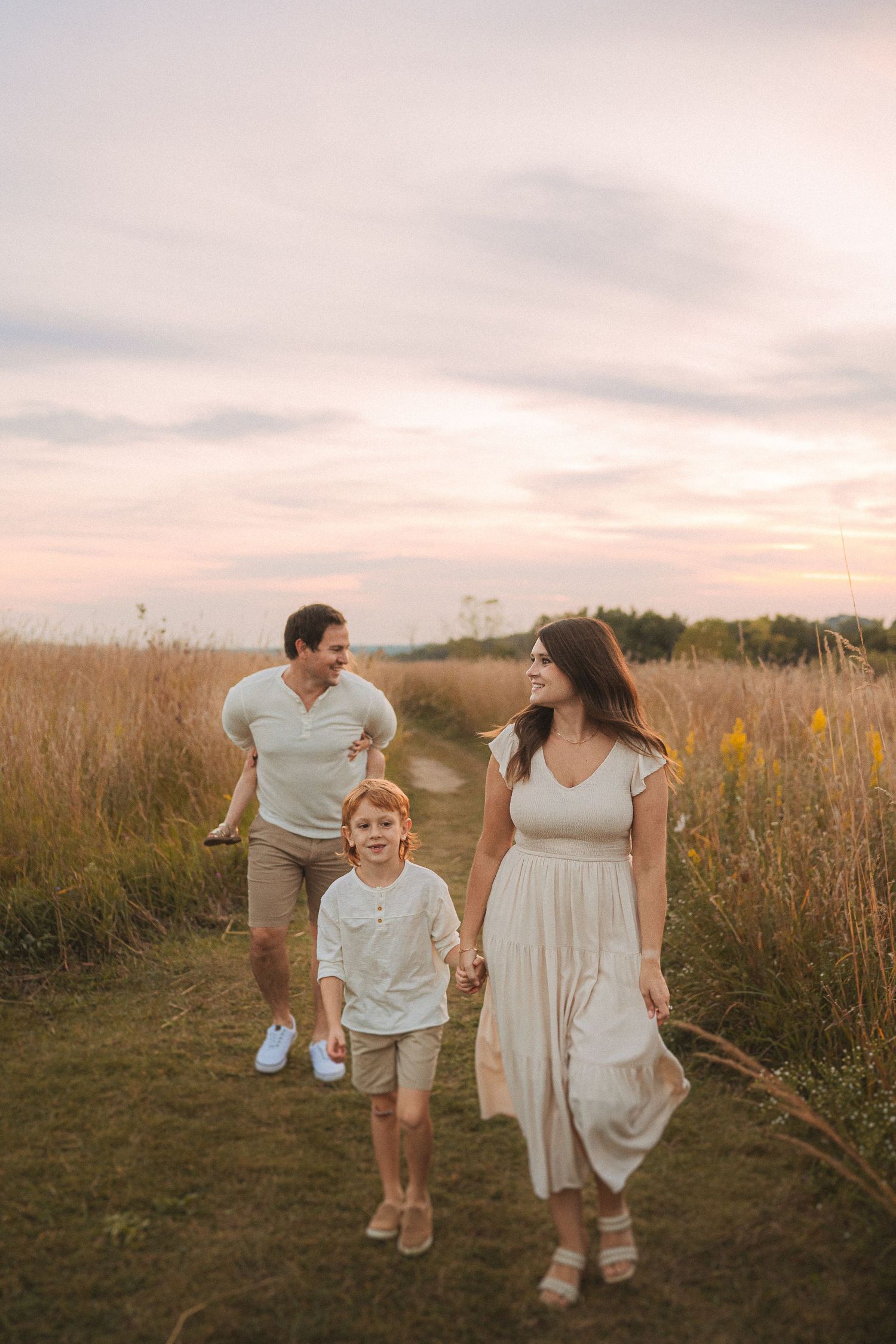Family walking together on a dirt path through tall grass during sunset.