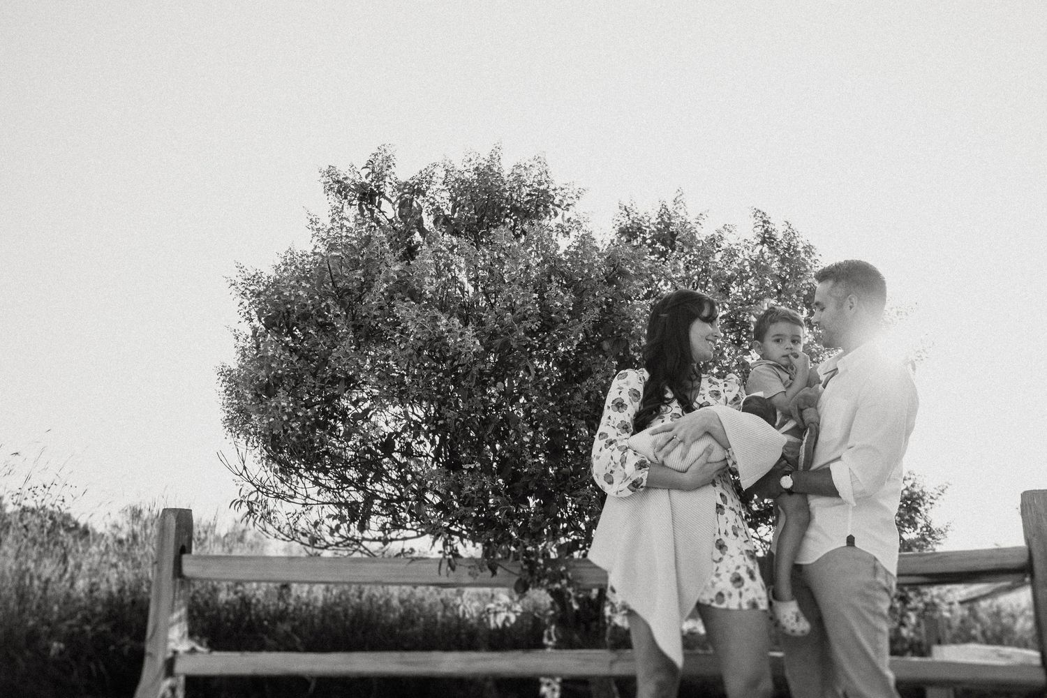 Couple stands by wooden fence near lush bush in black and white photo.
