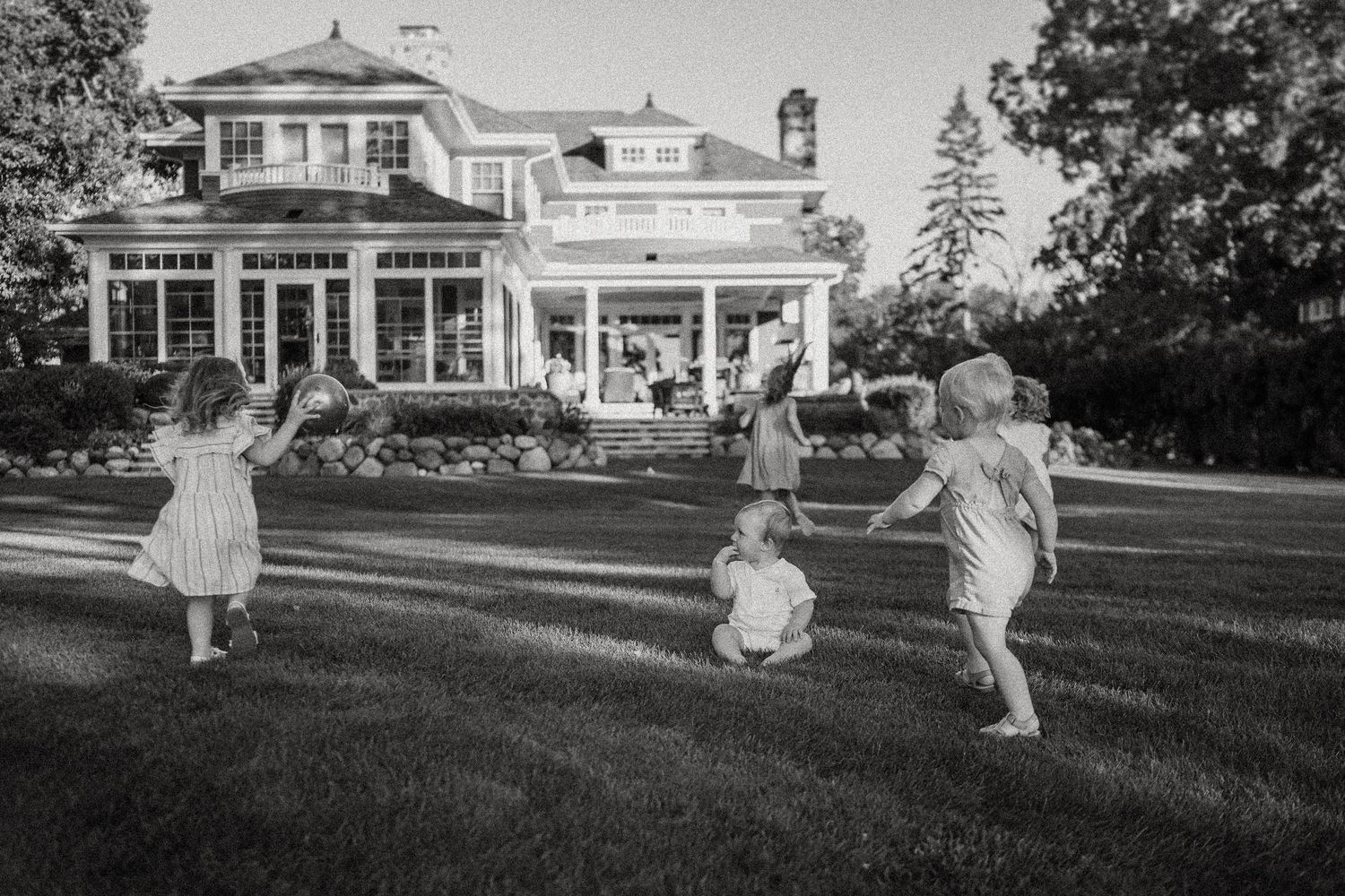 Children play and run on the lawn in front of a stately white mansion in black and white.