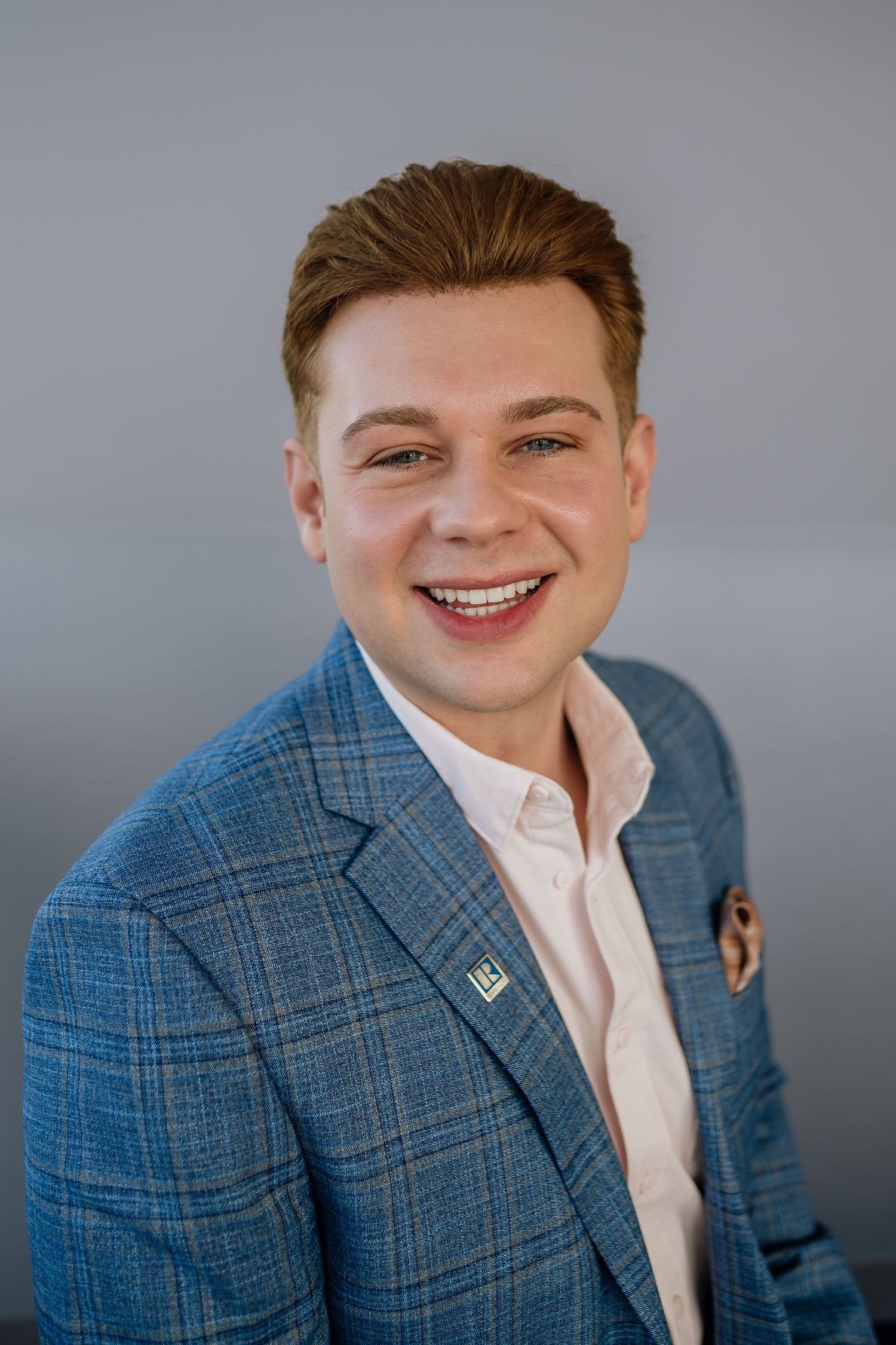Business headshot showing blue checkered blazer with pocket square against neutral background.