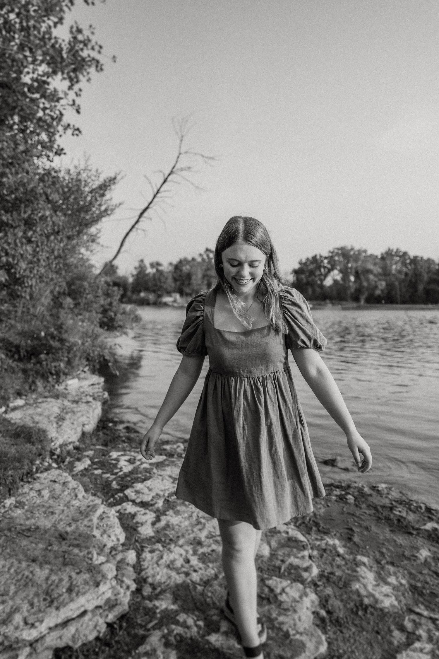 A black and white image of someone walking along rocky lakeside terrain in a flowing dress.