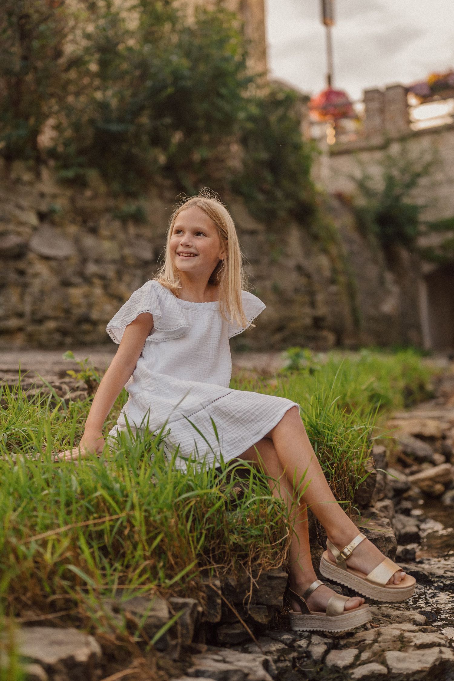 Child in white dress sits on rocky ground with tall grass and stone wall in background.