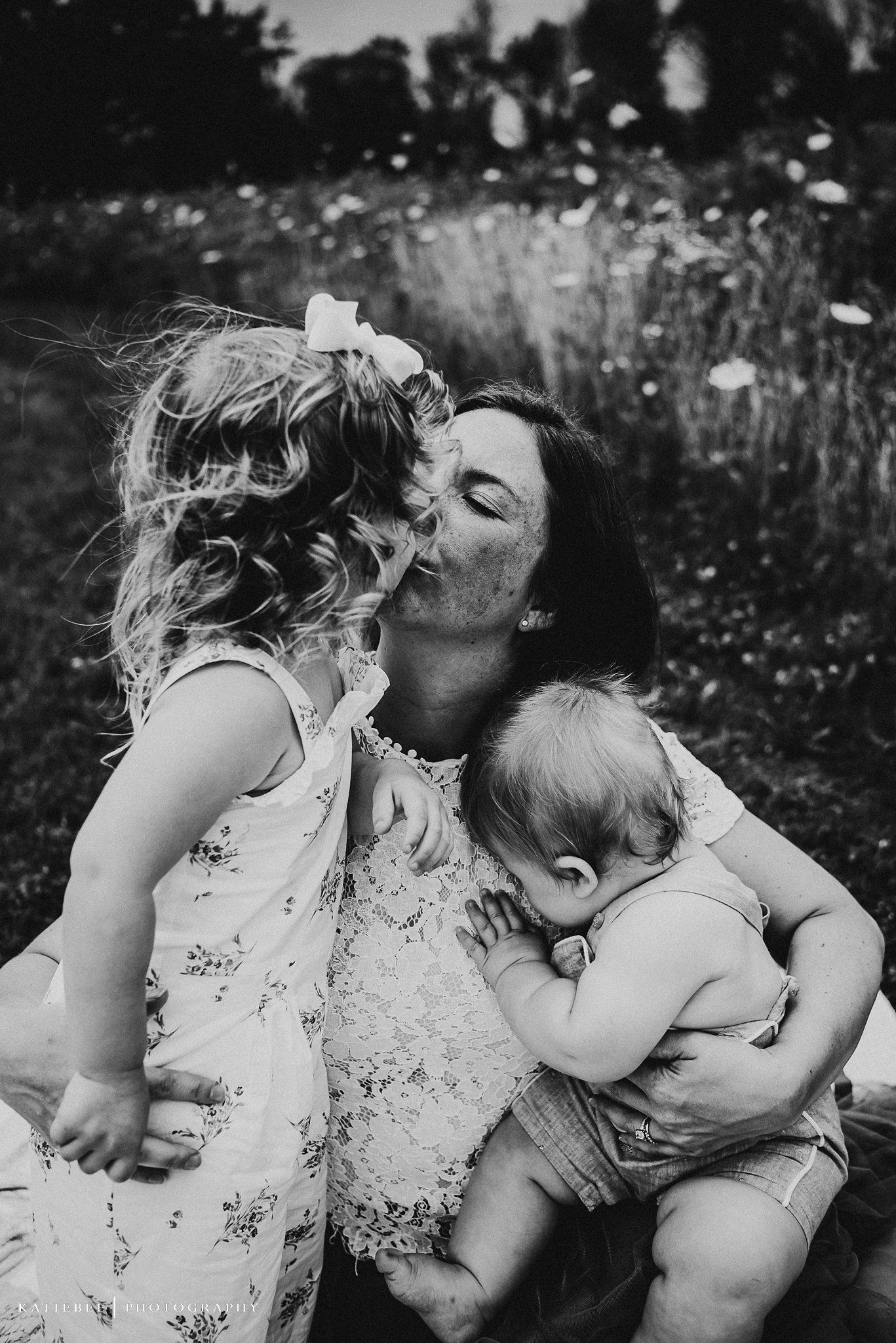Tender family moment captured in black and white as mother embraces two small children in a wildflower meadow.