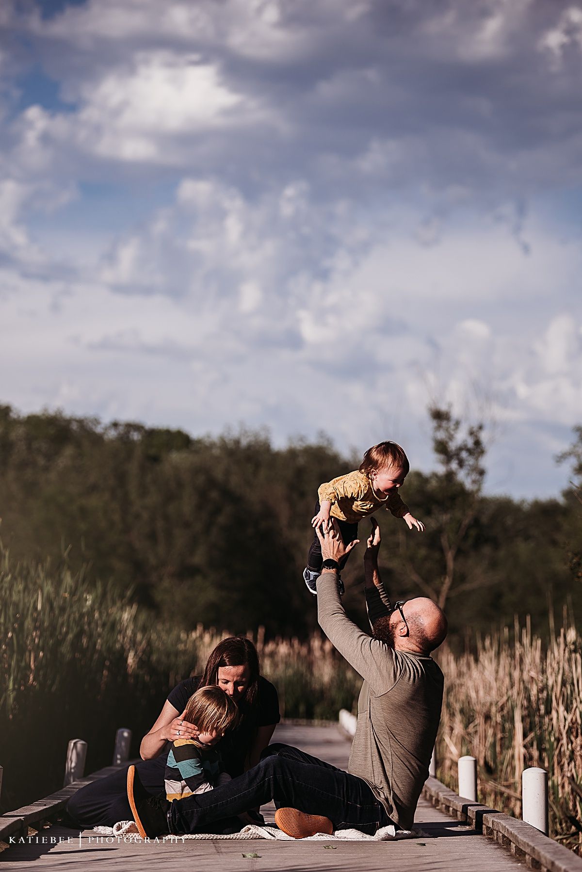Playful outdoor family moment captured on wooden boardwalk during golden hour.