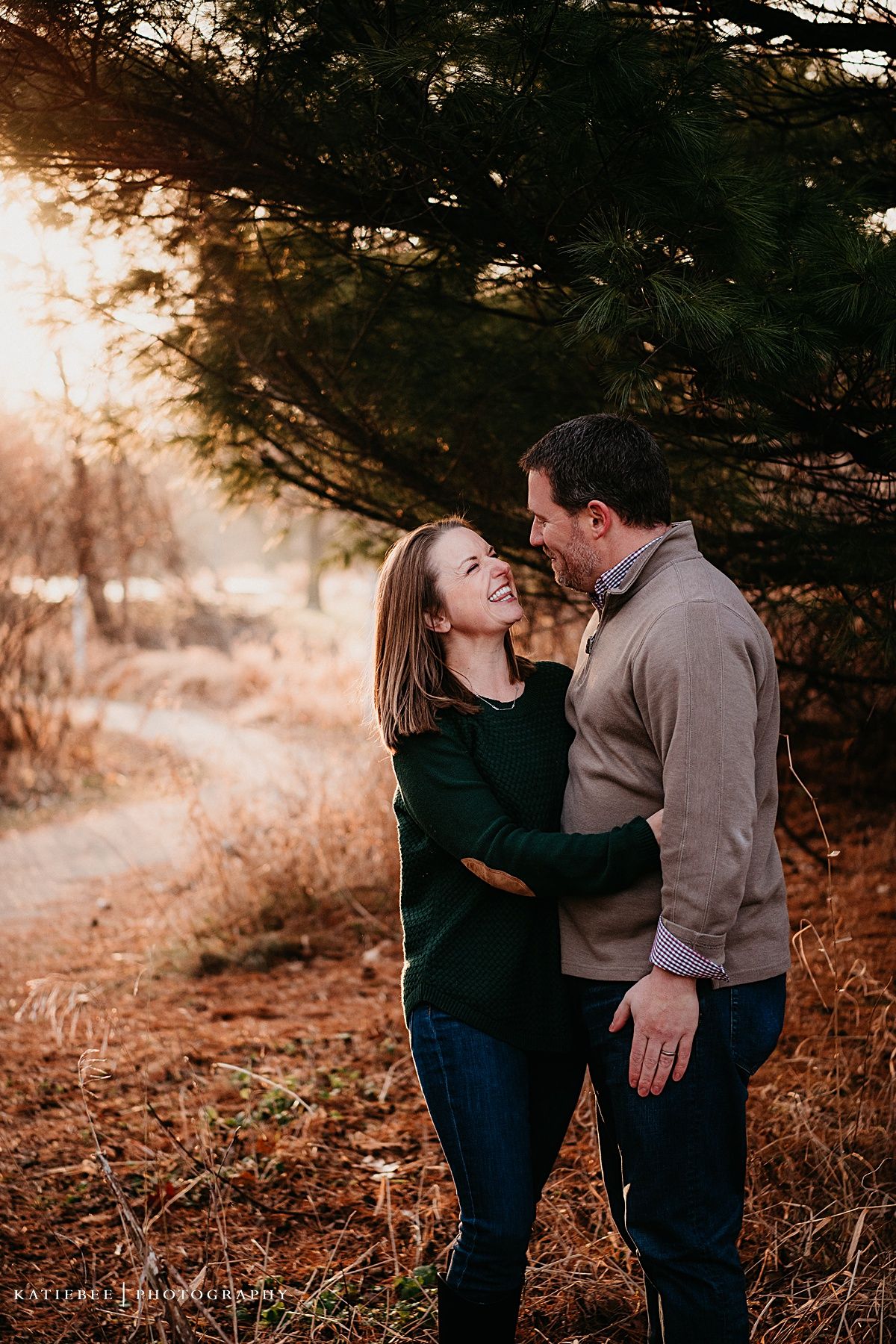 A couple shares an intimate moment while laughing together in a forest setting at sunset.