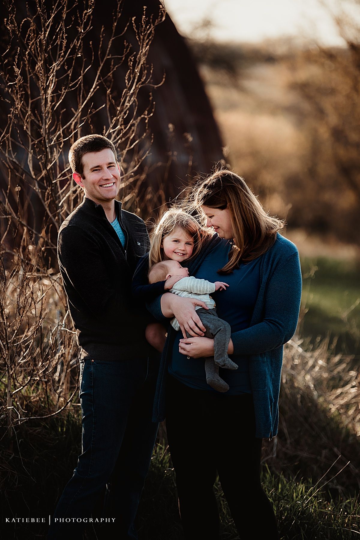 Loving family portrait captured during golden hour against rustic outdoor backdrop.