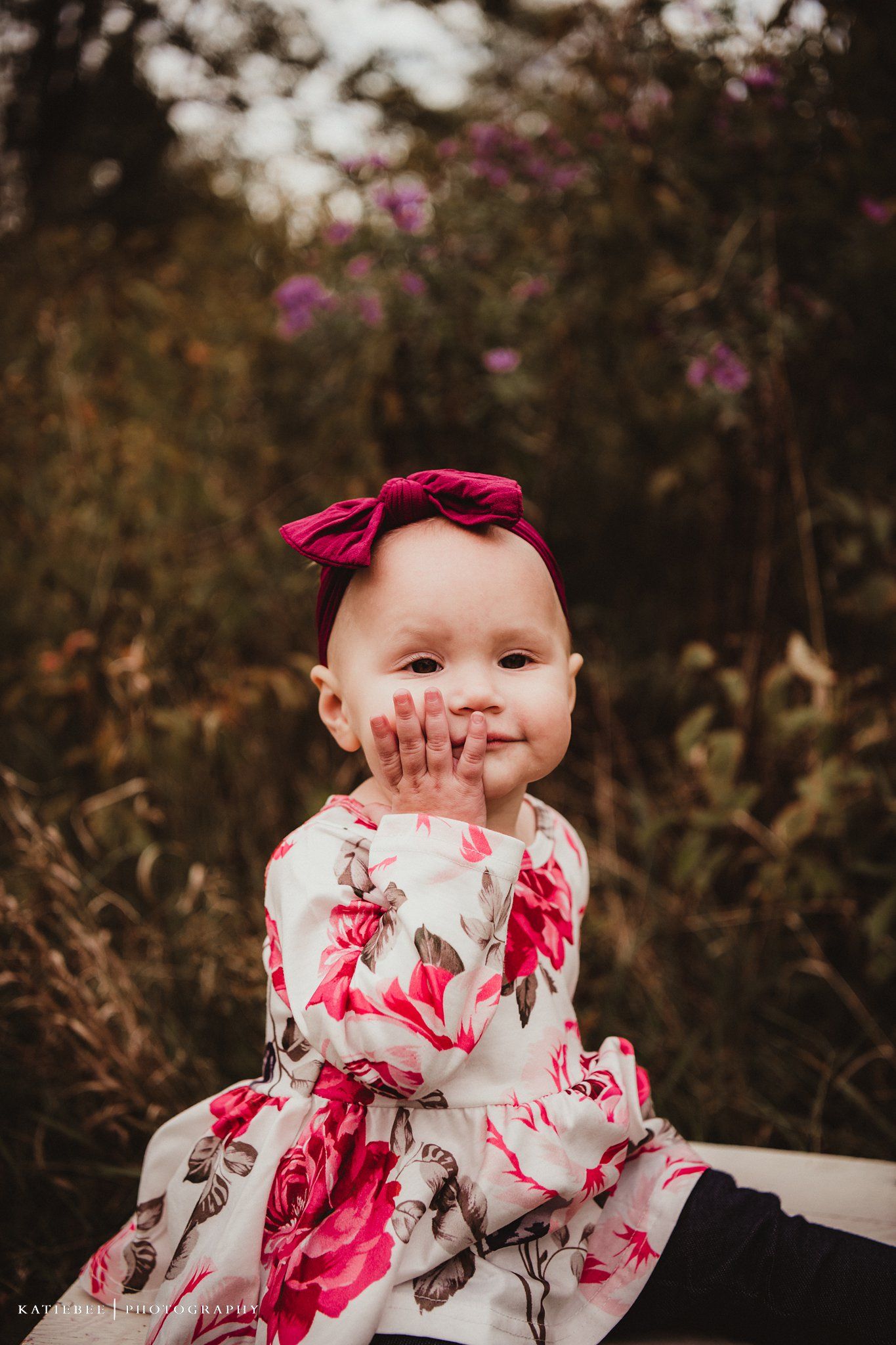 Baby in floral dress and red bow playfully covers mouth while smiling.