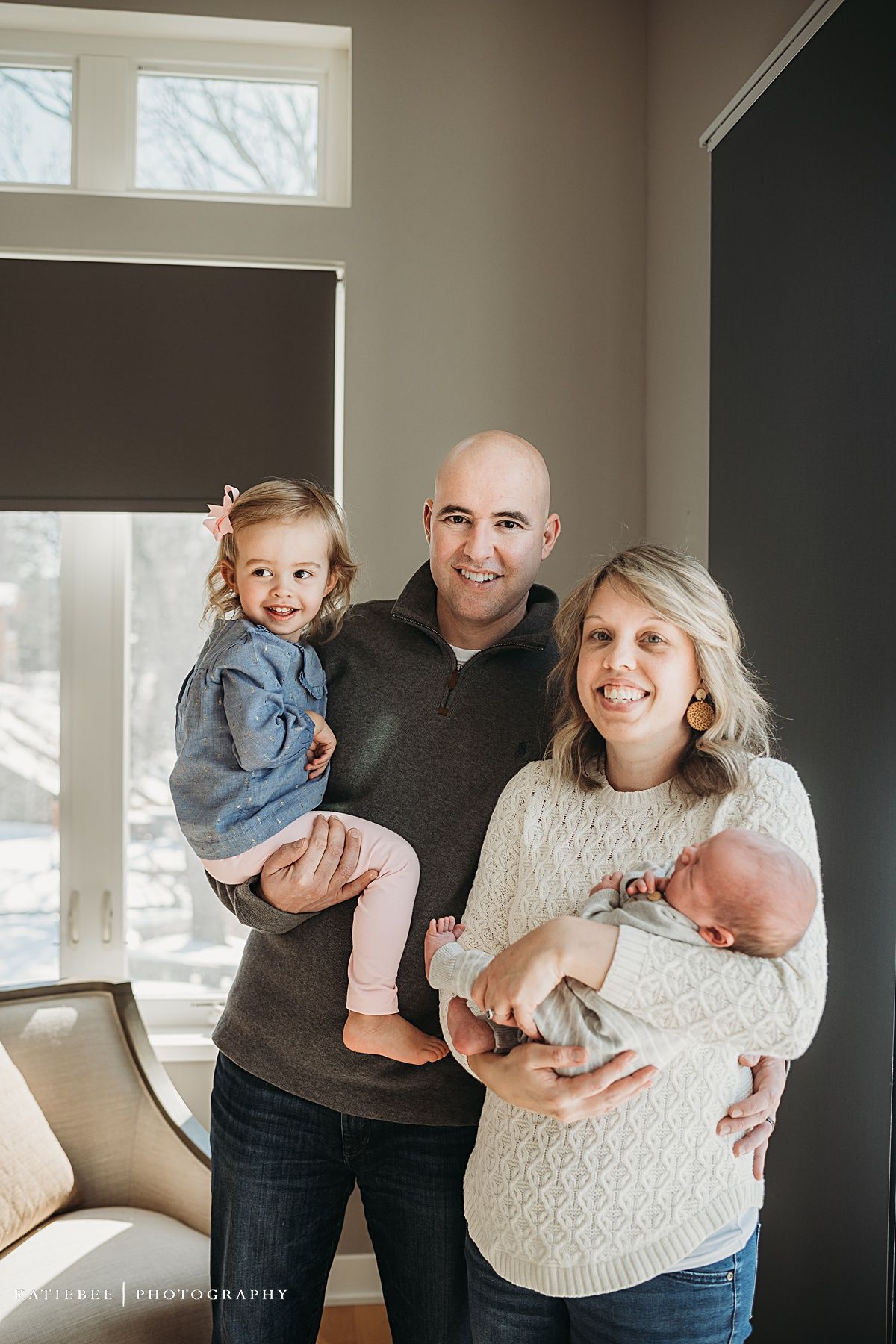 Natural light family portrait showing parents with their toddler and newborn in a cozy living room.