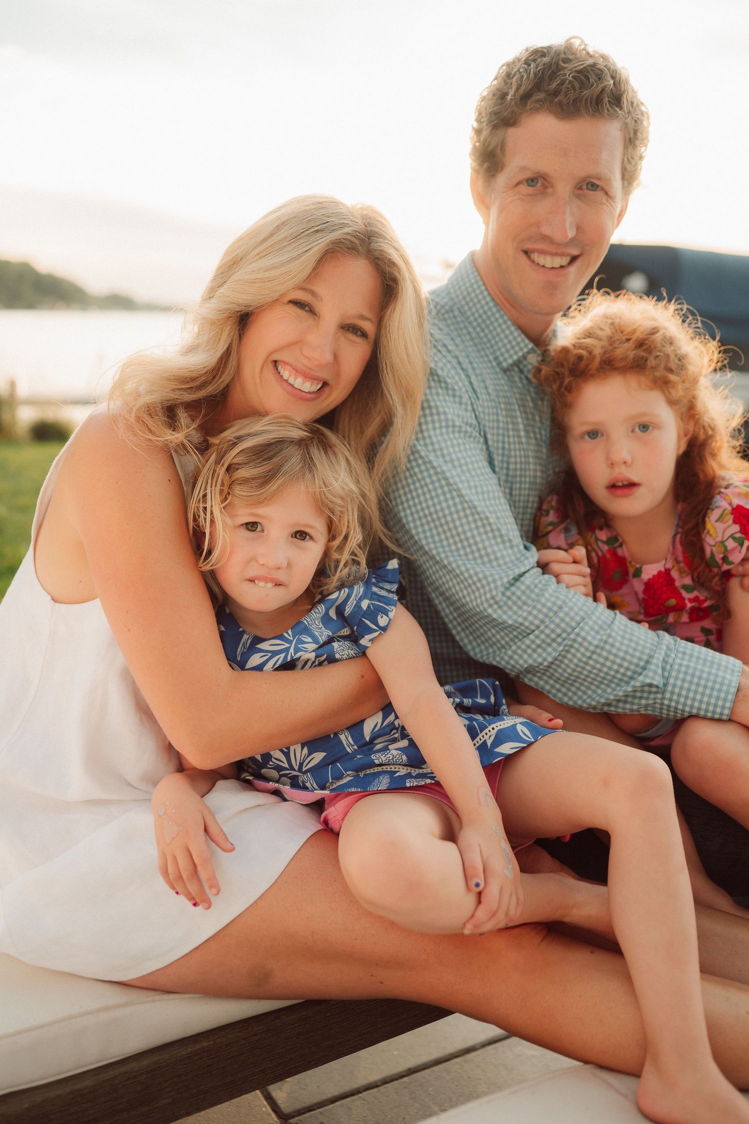 A family sits together on a boat deck at sunset, wearing casual summer attire with water and trees in the background.