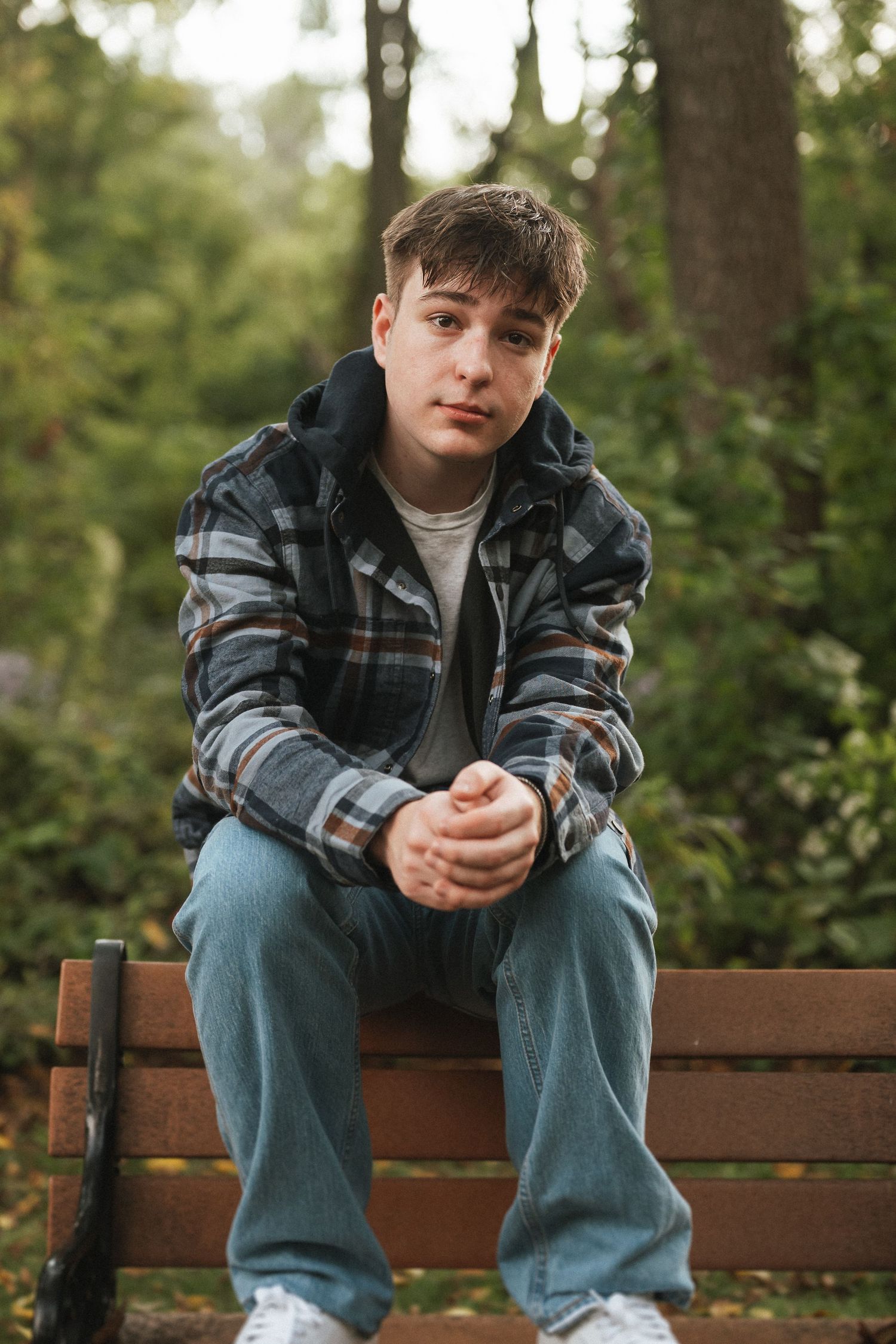 Someone sits on a wooden park bench wearing jeans and a plaid jacket while looking down at their hands.