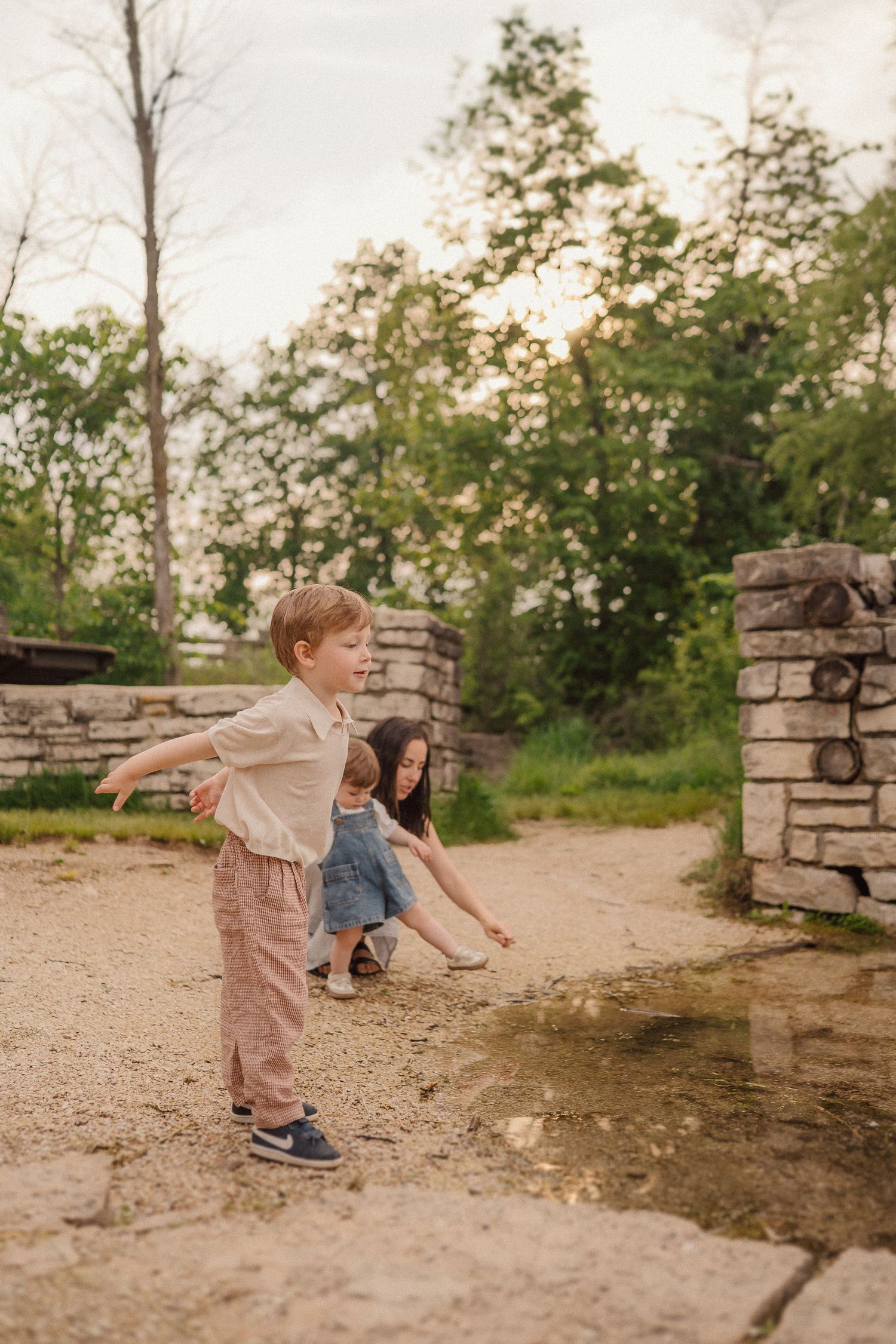 A toddler in casual clothes plays near a puddle by stone pillars at sunset in a natural setting.