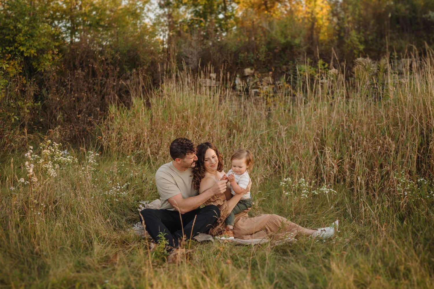 A family sits together on a blanket in a field of tall grass during sunset.