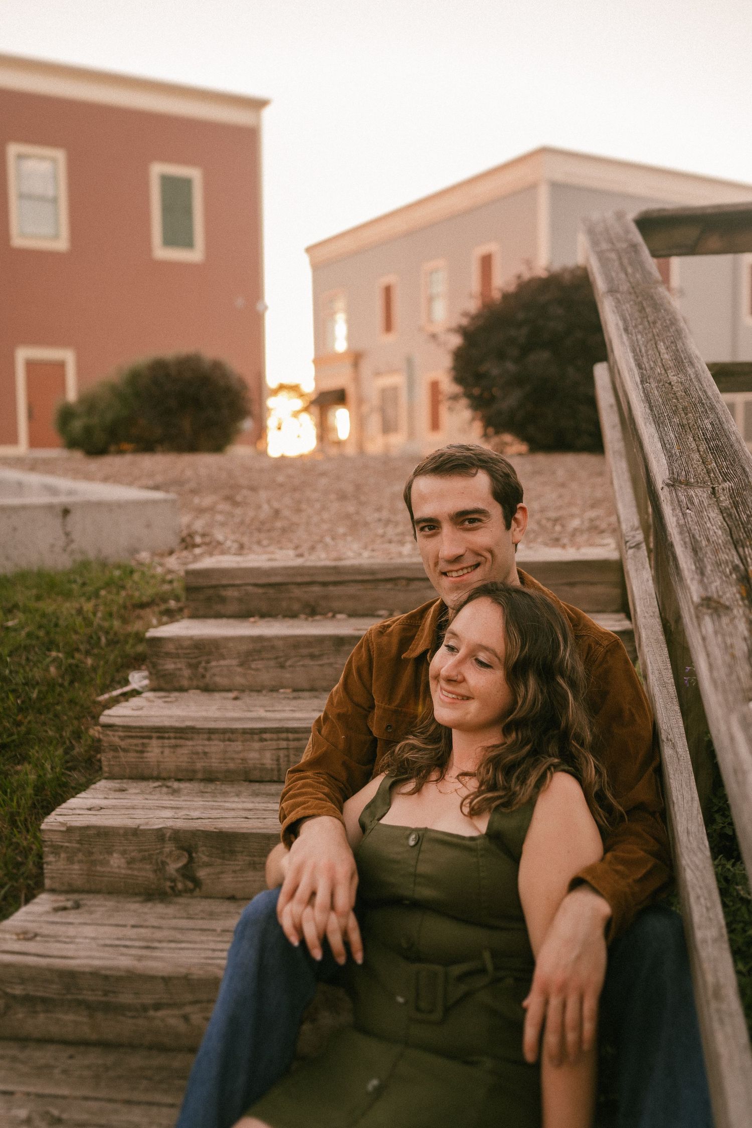 A couple sits together on outdoor wooden steps with pink buildings in background.