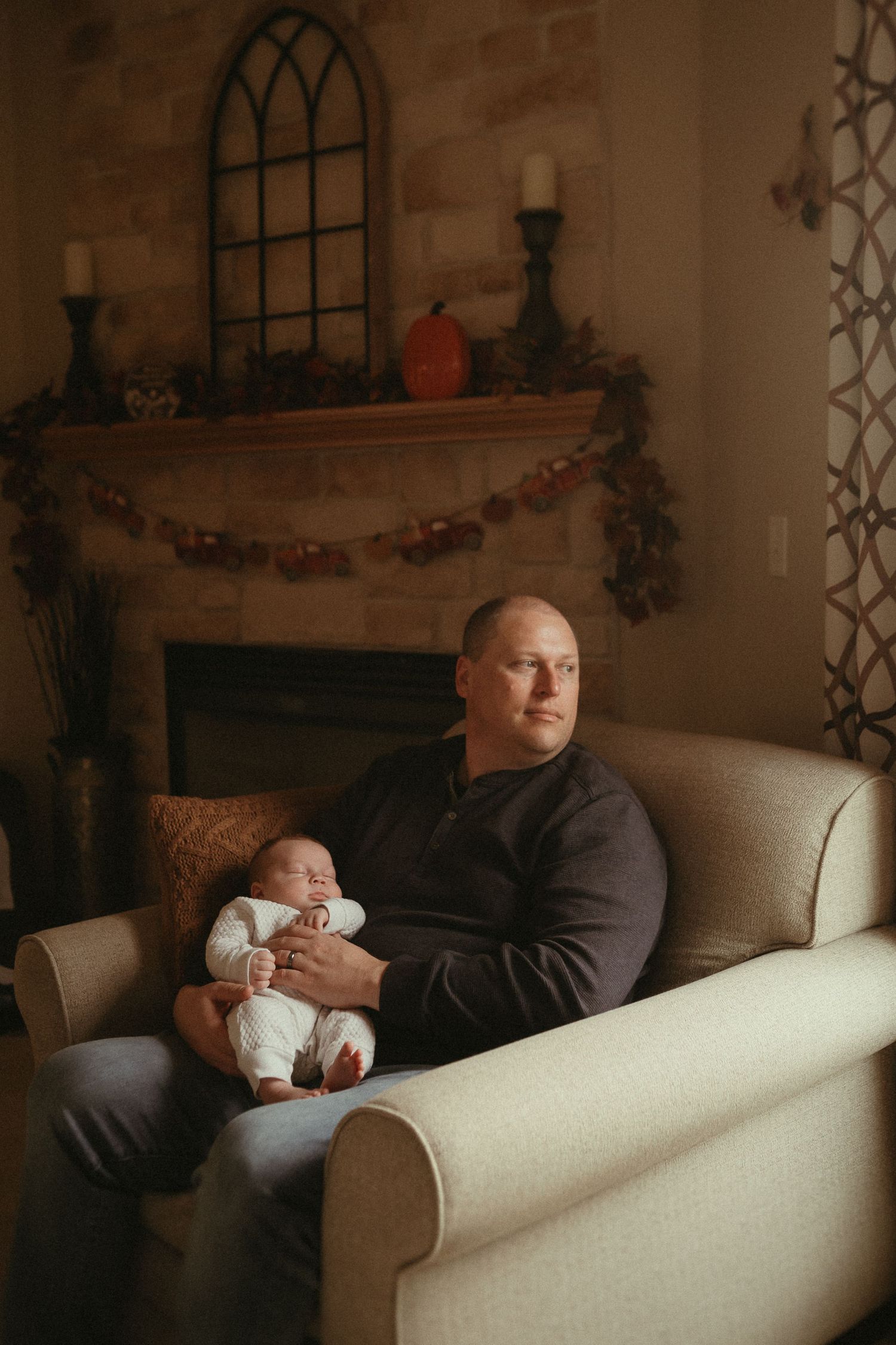 Cozy holiday scene with a parent and sleeping infant sitting on cream colored armchair by decorated fireplace mantle.