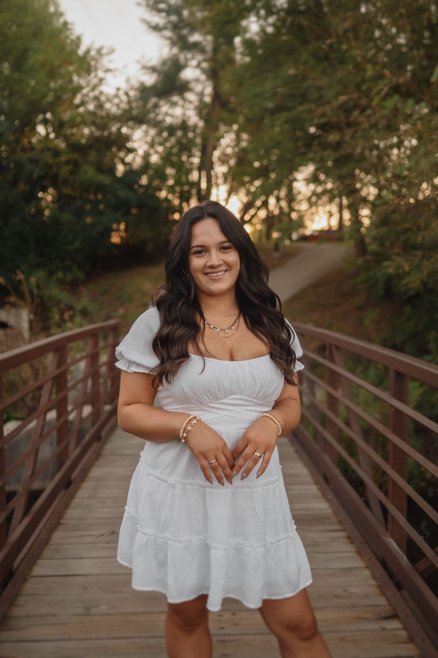 A woman in a white summer dress stands smiling on a wooden bridge path surrounded by trees at sunset.