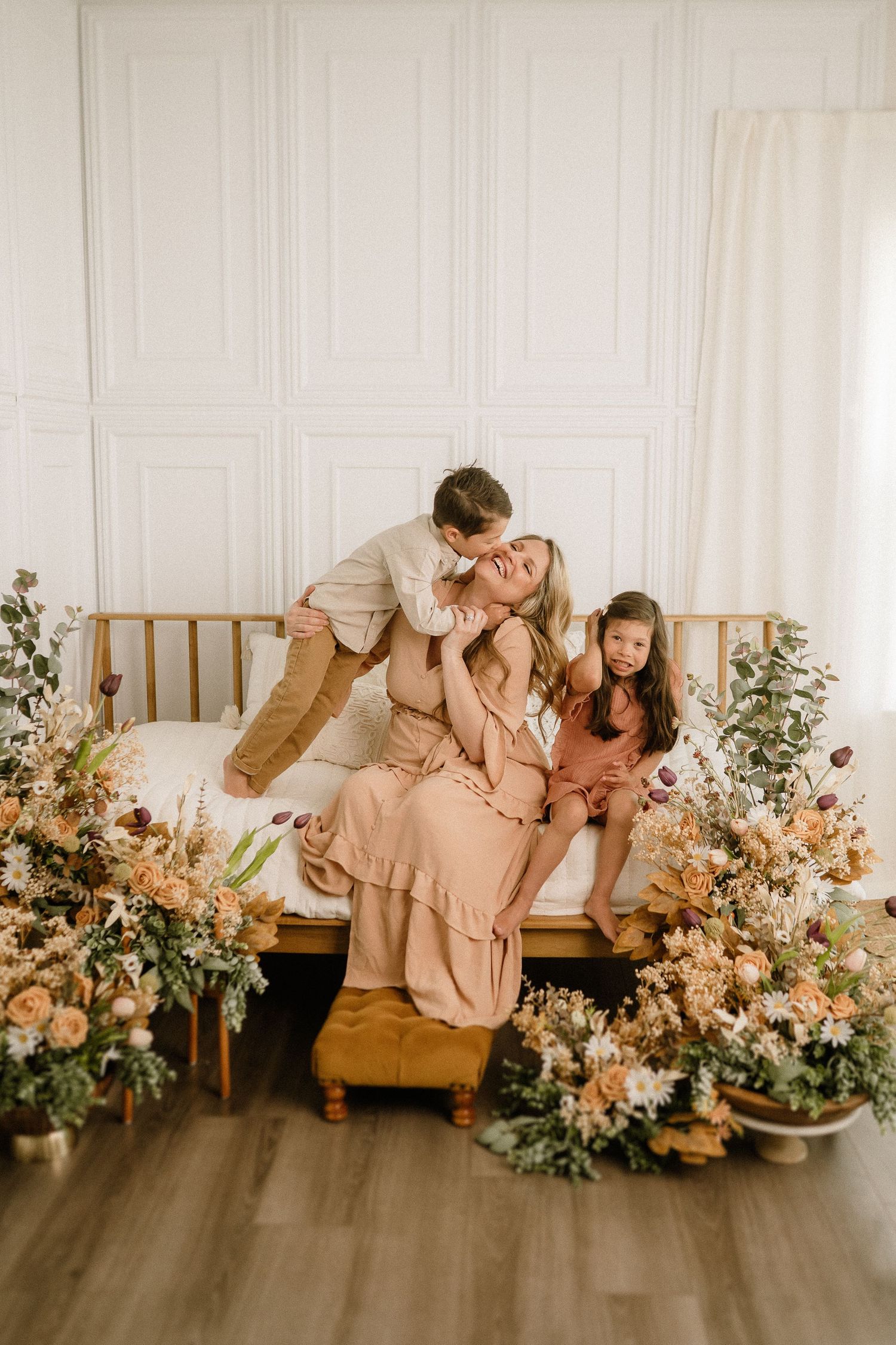 Family in neutral-toned outfits share tender moment on wooden bed surrounded by floral arrangements in bright room.
