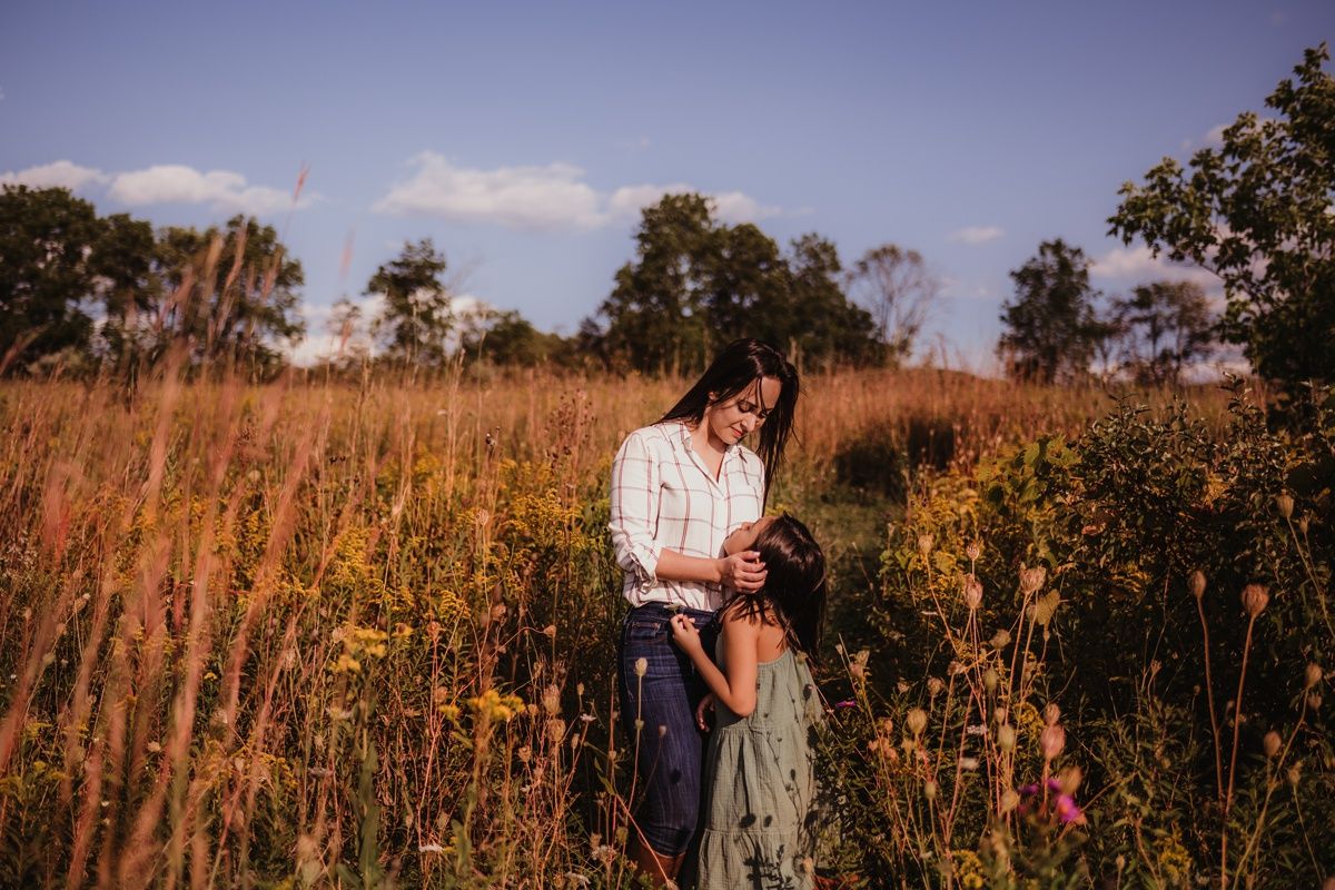 Two figures standing close together in a sunlit meadow filled with tall golden grass and wildflowers.