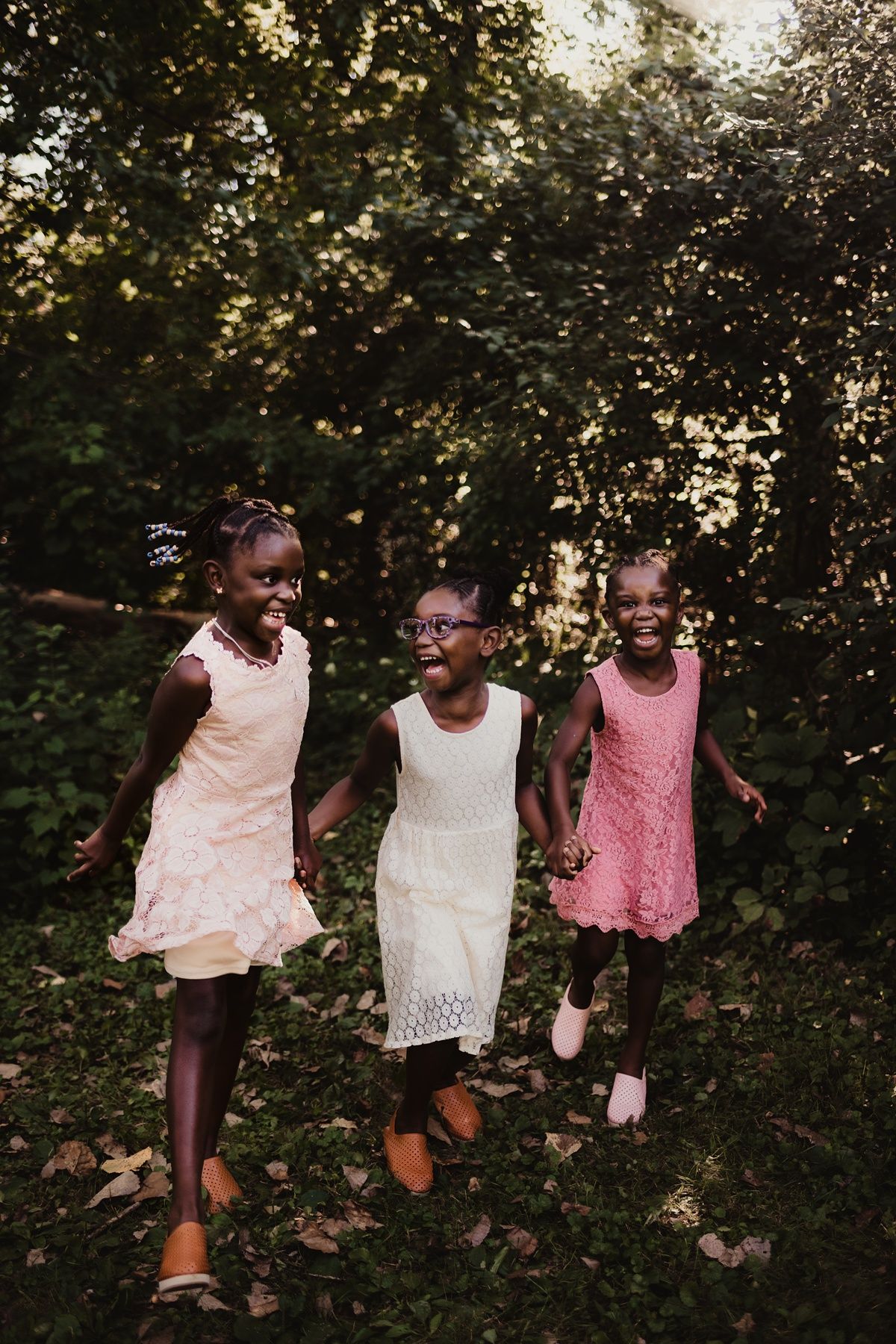 Three children in dresses holding hands while jumping and playing in a wooded area.