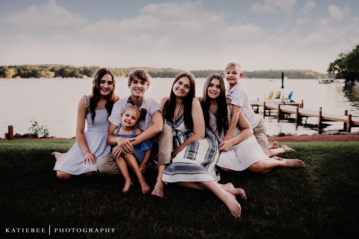 Group sitting together on grassy lakefront with dock and water in background.