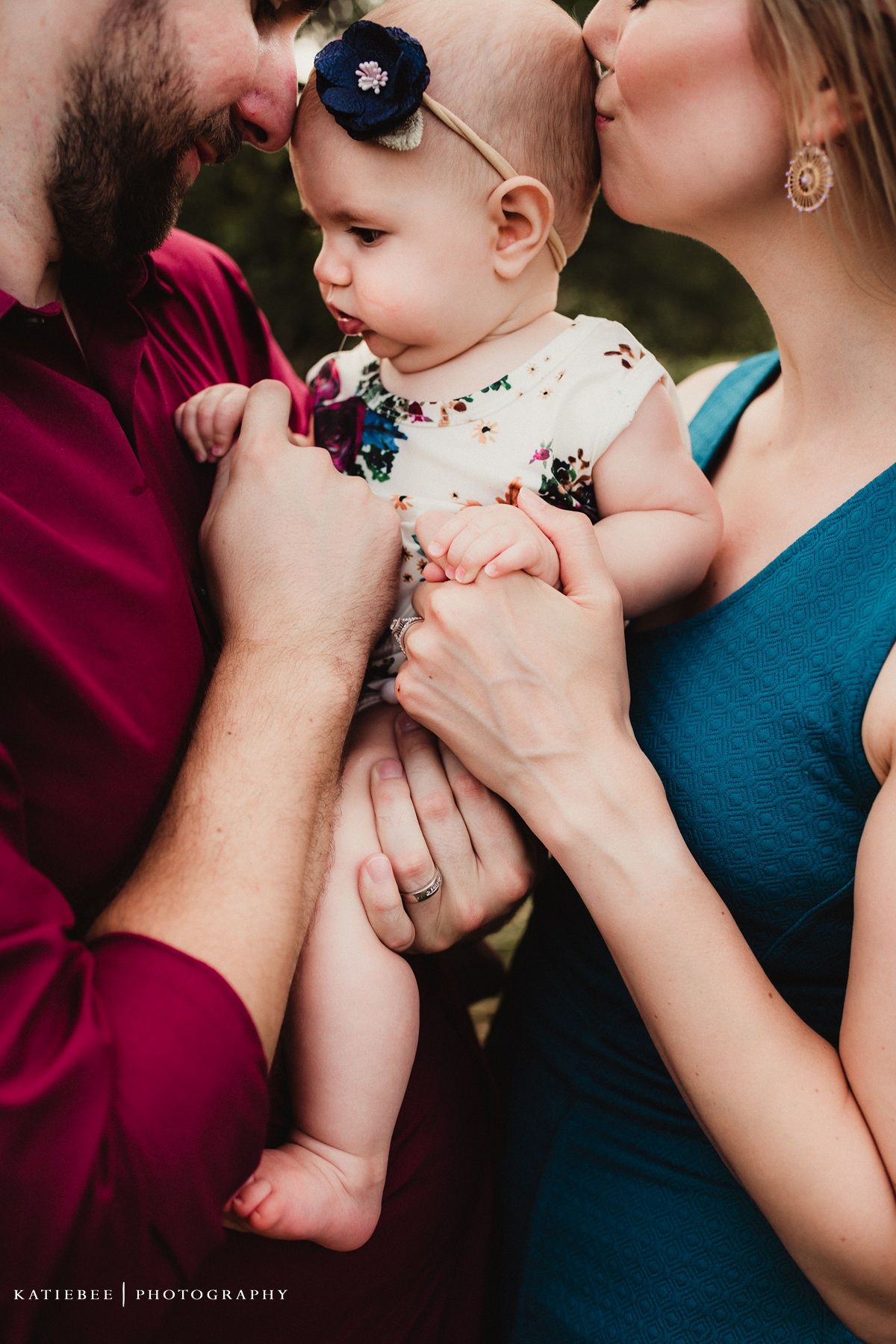Close up shot of a baby in a floral outfit with a navy bow headband being held and kissed.