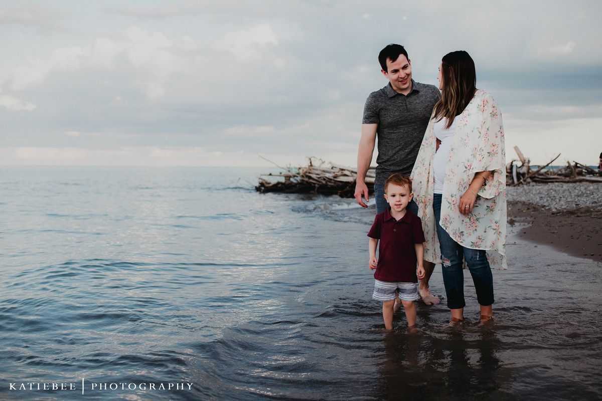 Family standing in shallow water at sunset on a rocky beach with driftwood visible.