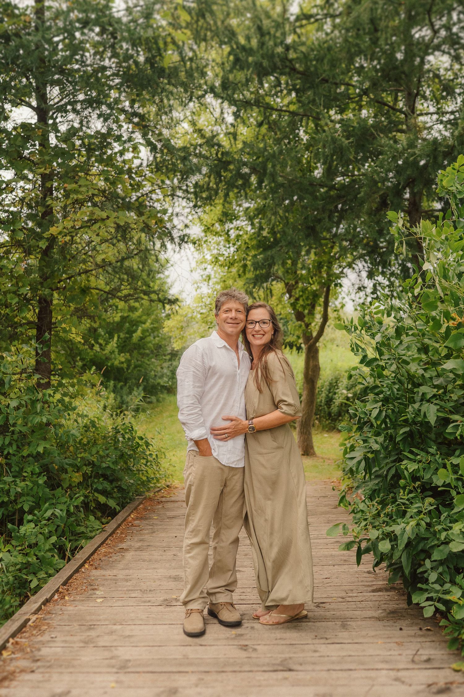 A couple poses together on a wooden boardwalk surrounded by green trees and natural lighting.