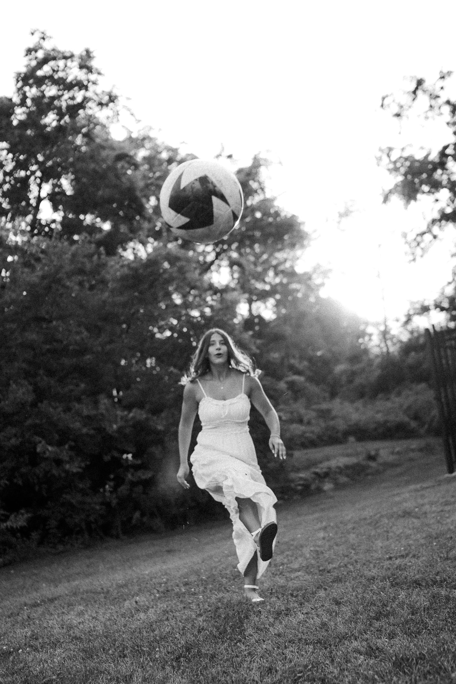 A person in a white dress jumps playfully with a volleyball against a sunset sky.