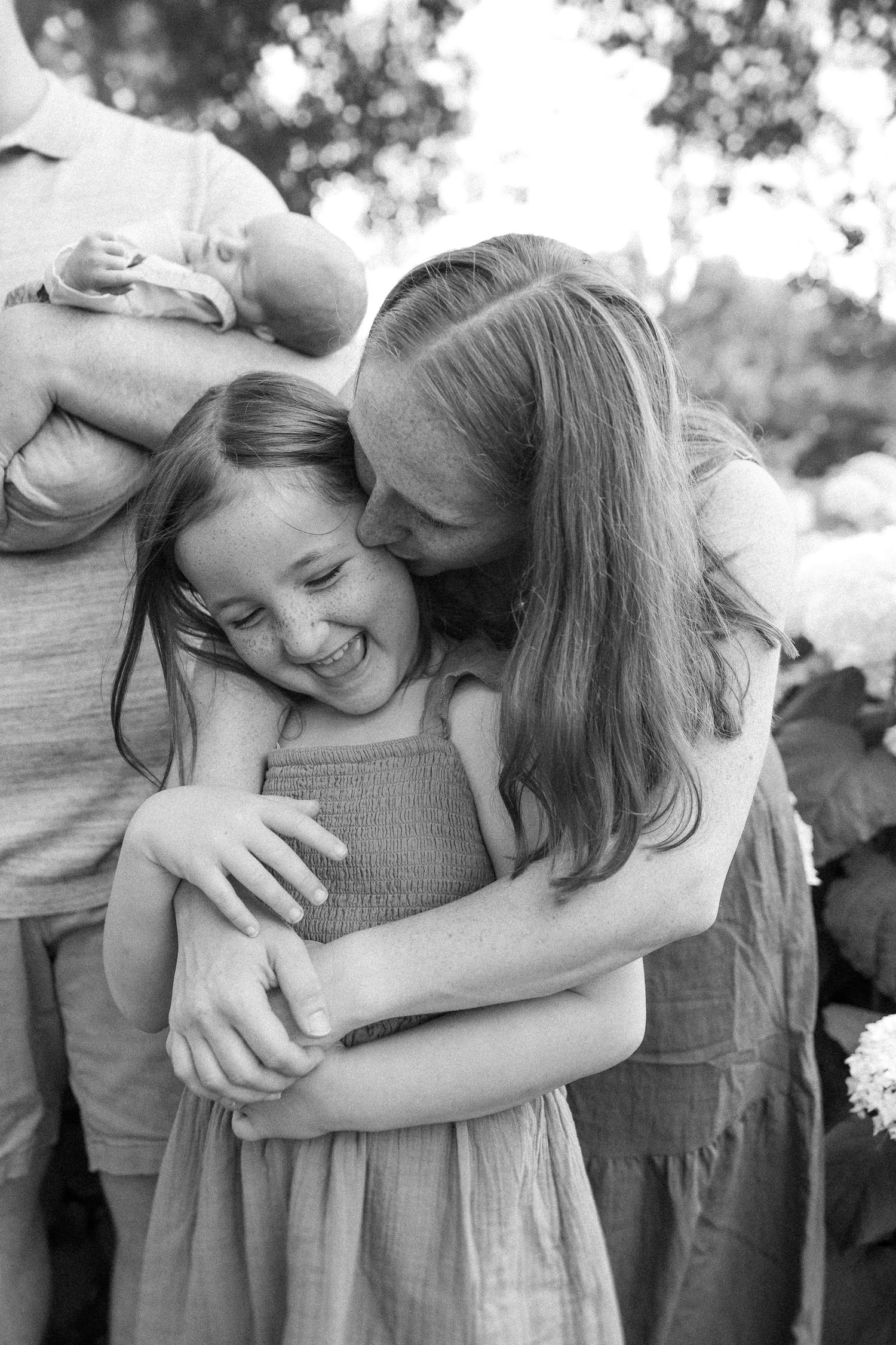 Black and white candid photo of an intimate playful moment with warm hugs and genuine laughter in an outdoor setting.