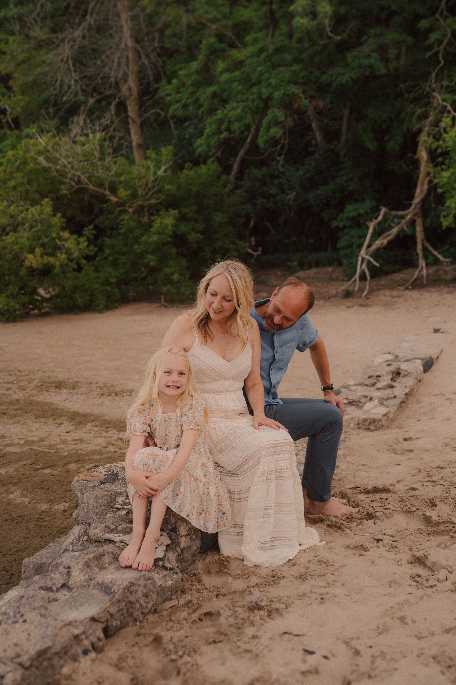 A family sits together on a sandy beach shore against a backdrop of lush green forest trees at sunset.
