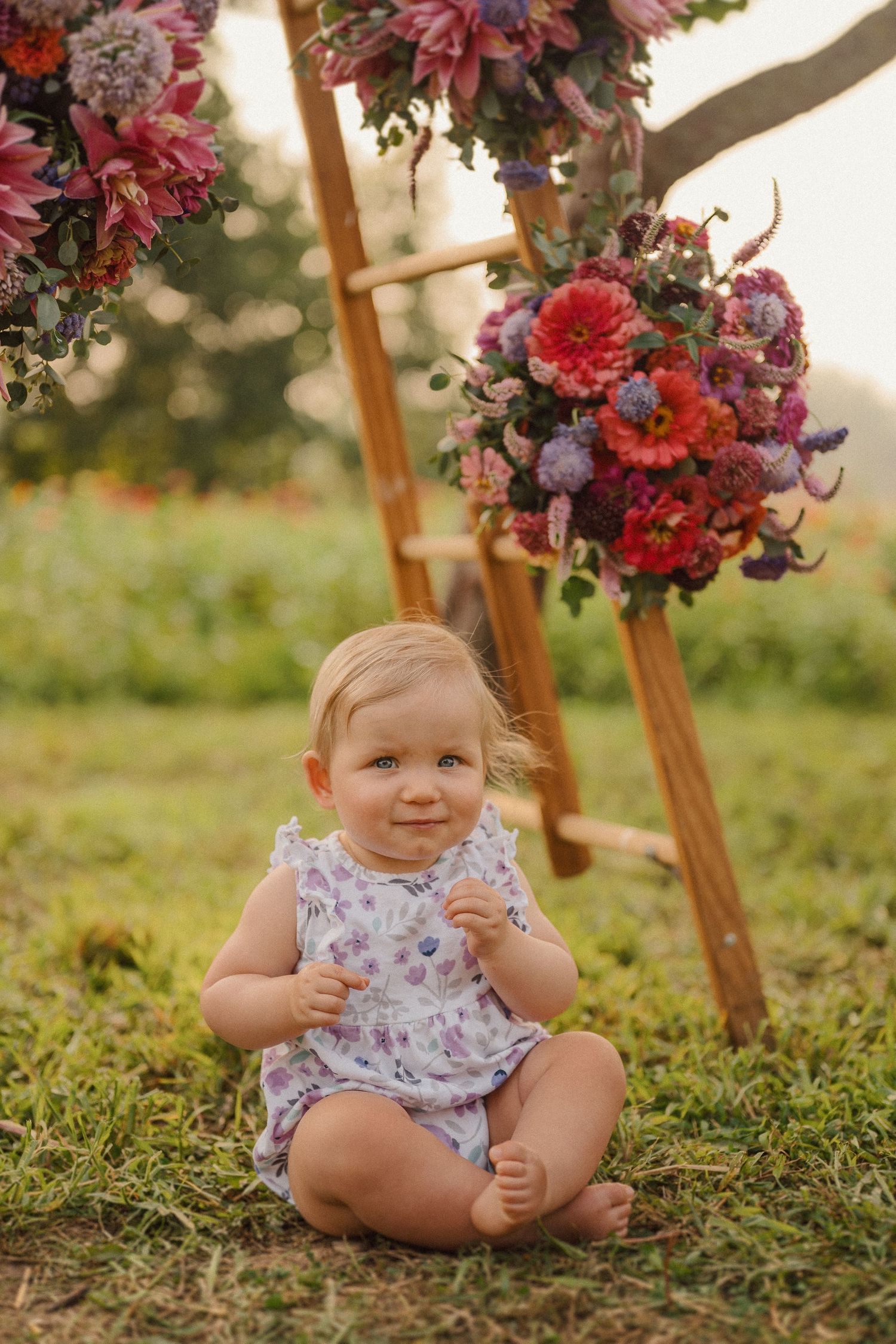 A baby sits on grass beneath a decorative ladder adorned with pink and purple flowers in a garden setting.