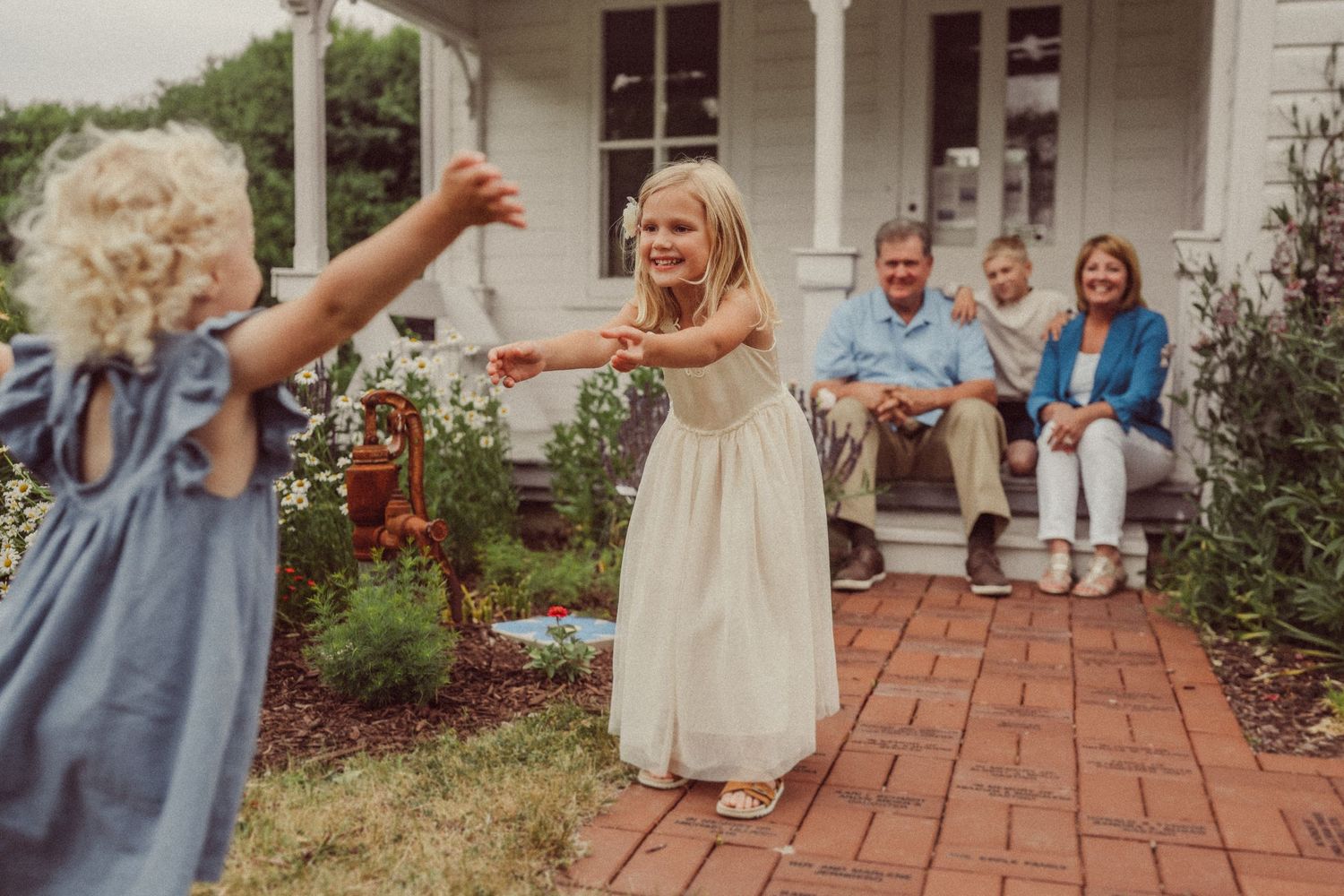 Children play on a brick patio while adults watch from white porch steps.