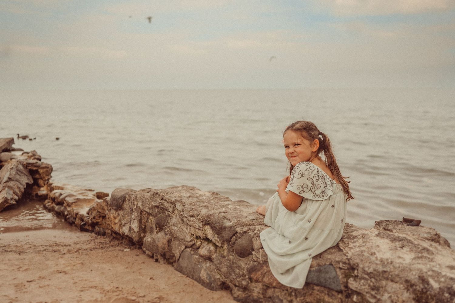 A person in a white dress sits contemplatively on rocky beach at sunset overlooking the calm ocean waters.