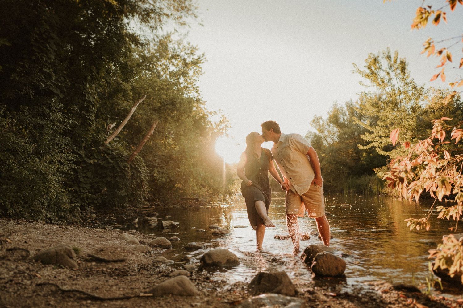 Two people stand together on rocks by a sunlit stream in a wooded area.