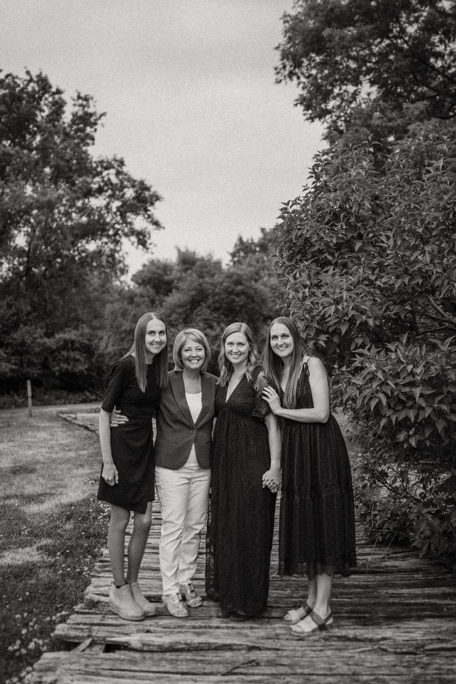 Four women standing together on wooden boardwalk surrounded by lush greenery in black and white.