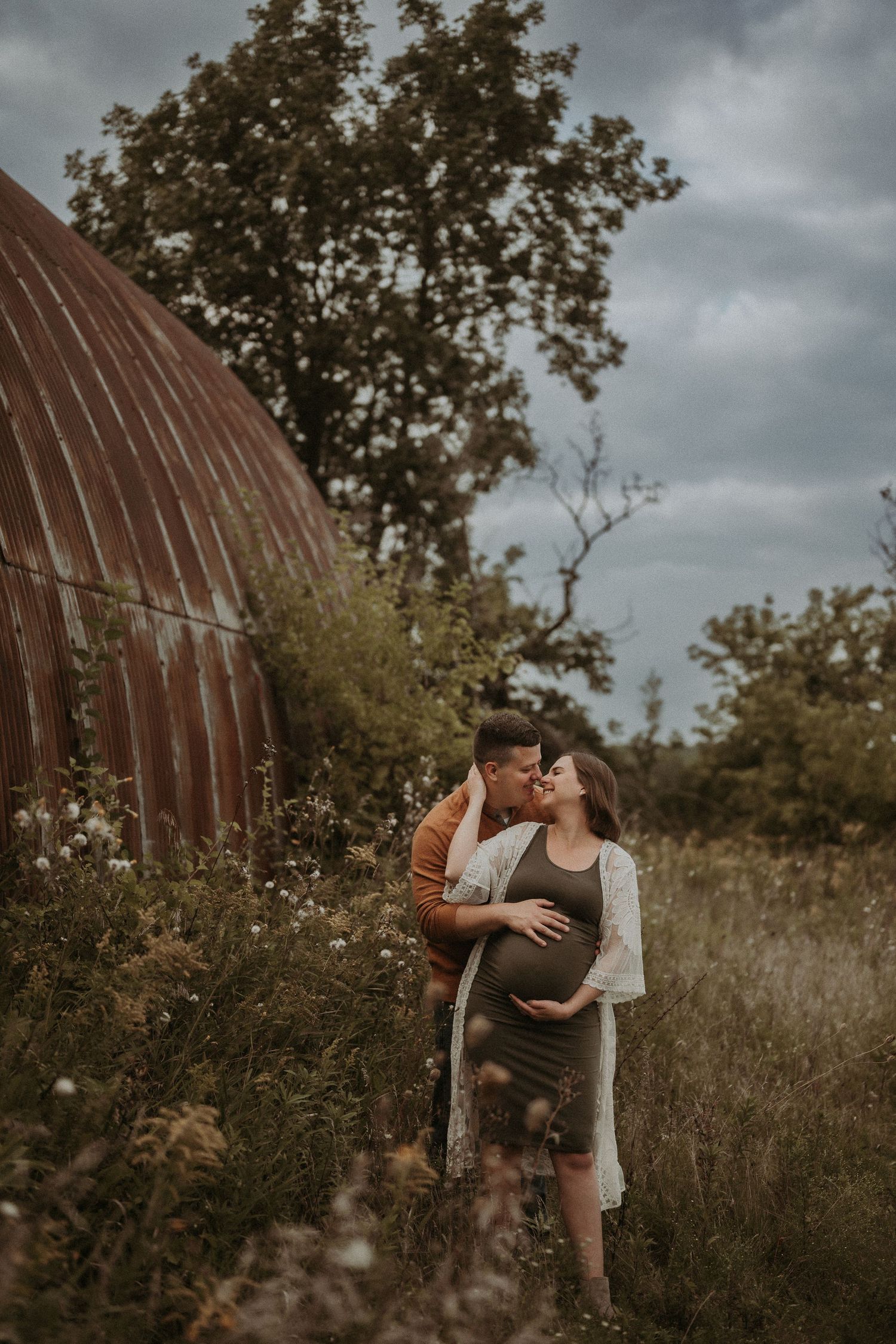 A romantic maternity photoshoot near a rustic barn structure at dusk.