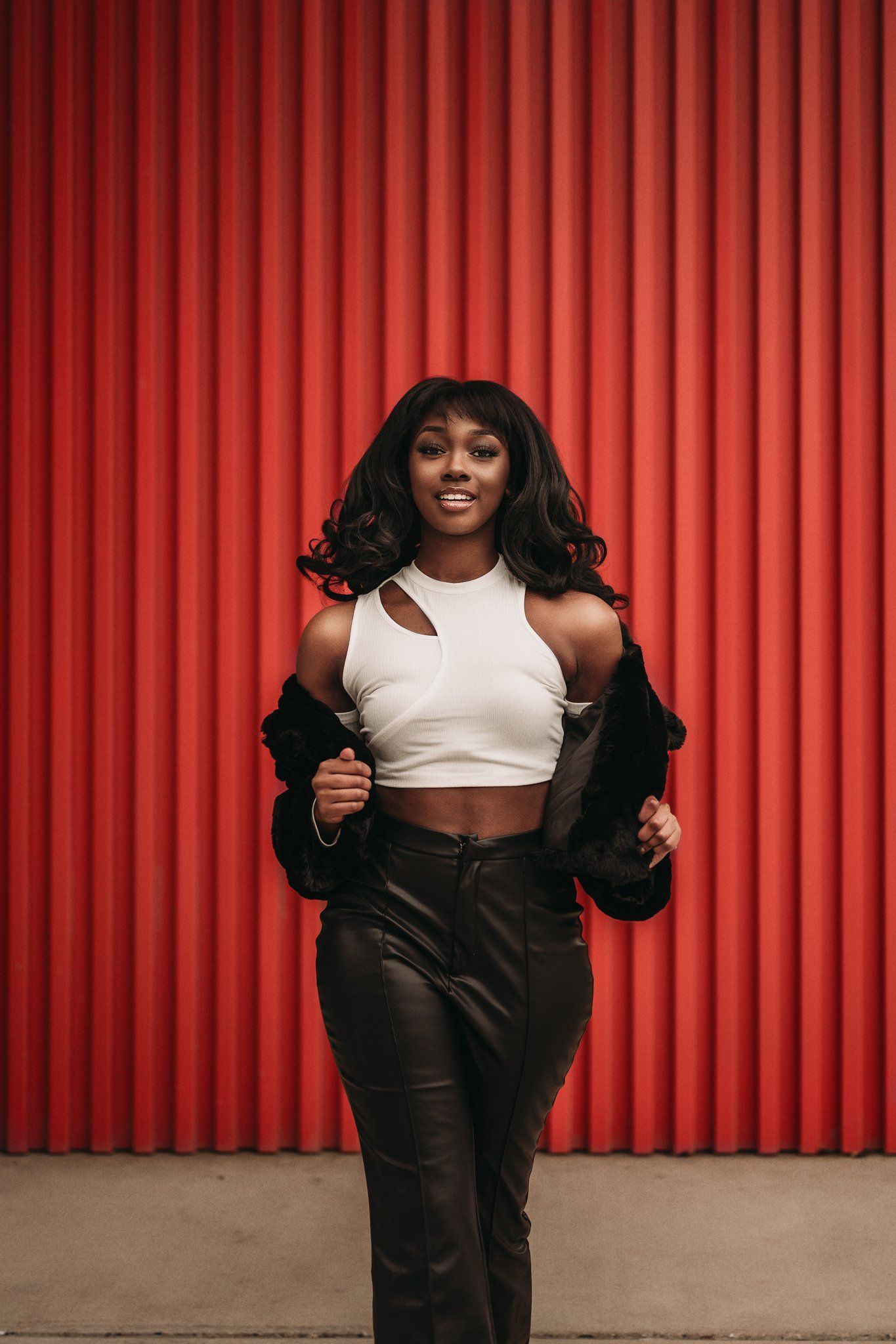 A fashion portrait featuring a model in a white crop top and black fur jacket against red stripes.
