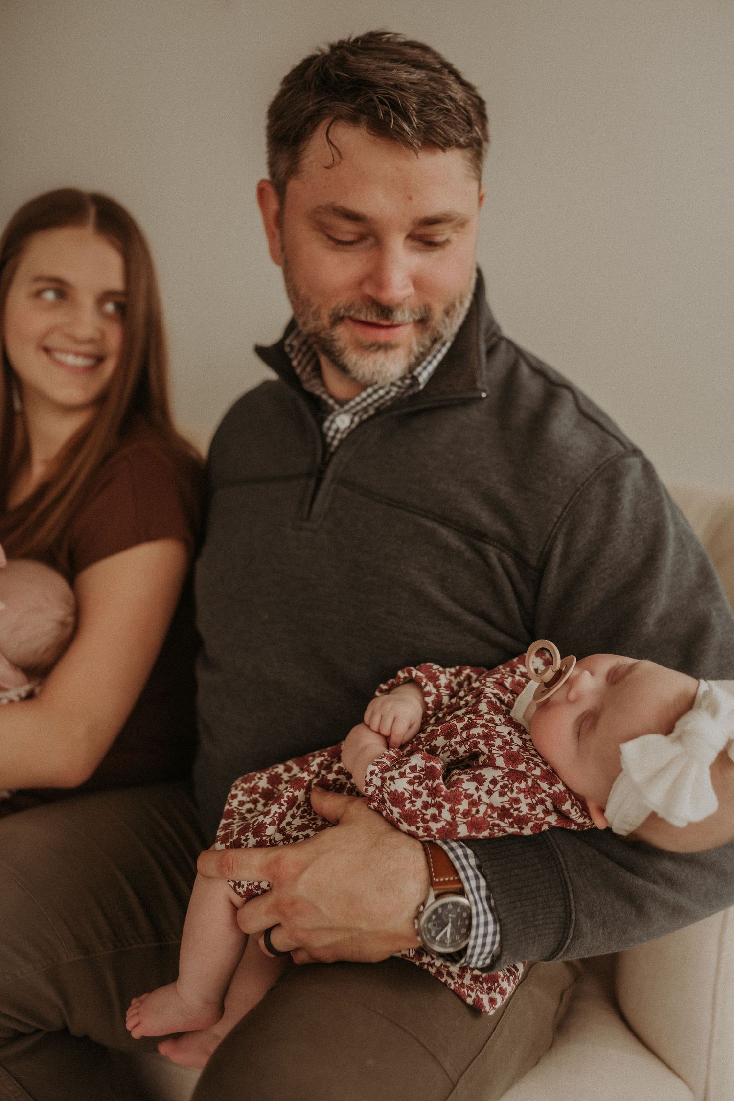 Parents share an intimate moment with their newborn baby in floral dress on a cream colored couch.
