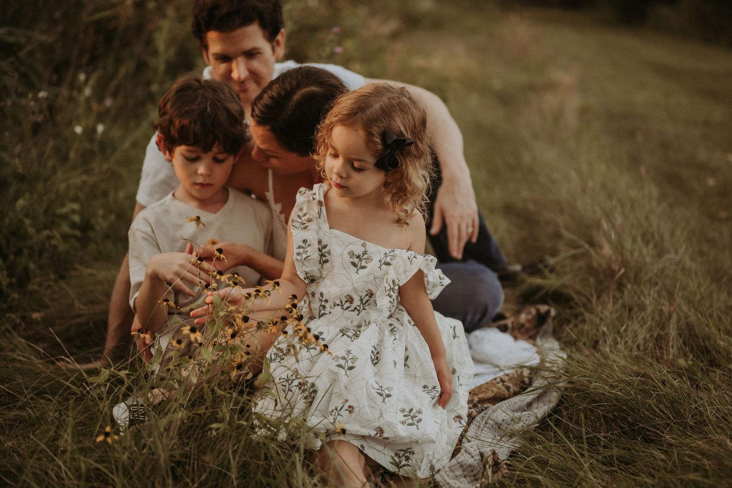 A family sits together in a field at sunset, with the young girl wearing a floral white dress.