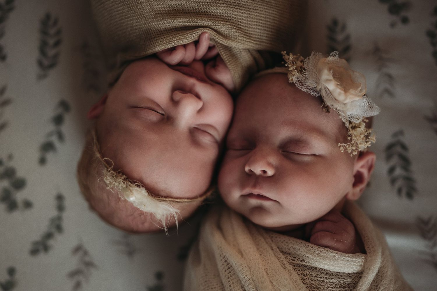 Sleeping newborn twins wearing delicate headpieces on floral bedding in close-up portrait.
