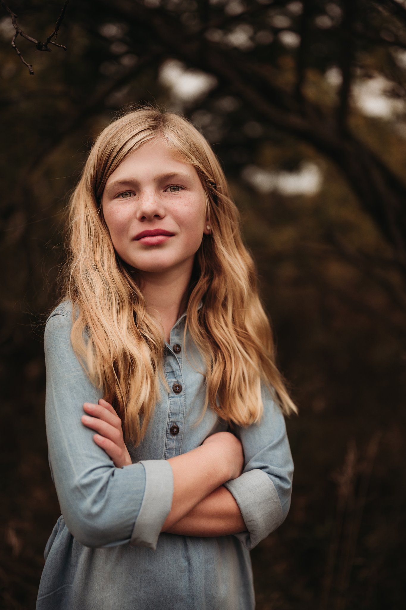 Person with long blonde hair wearing denim shirt poses with crossed arms and subtle smile outdoors.