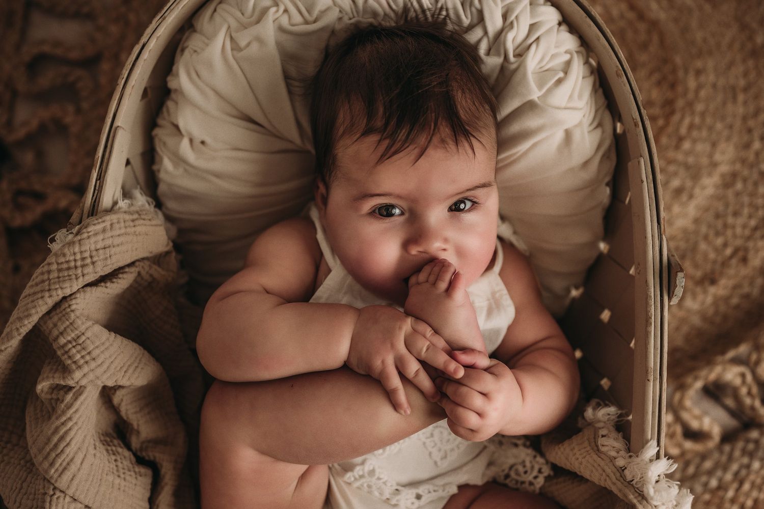 Baby holding white plush toy while resting in vintage cream bassinet.