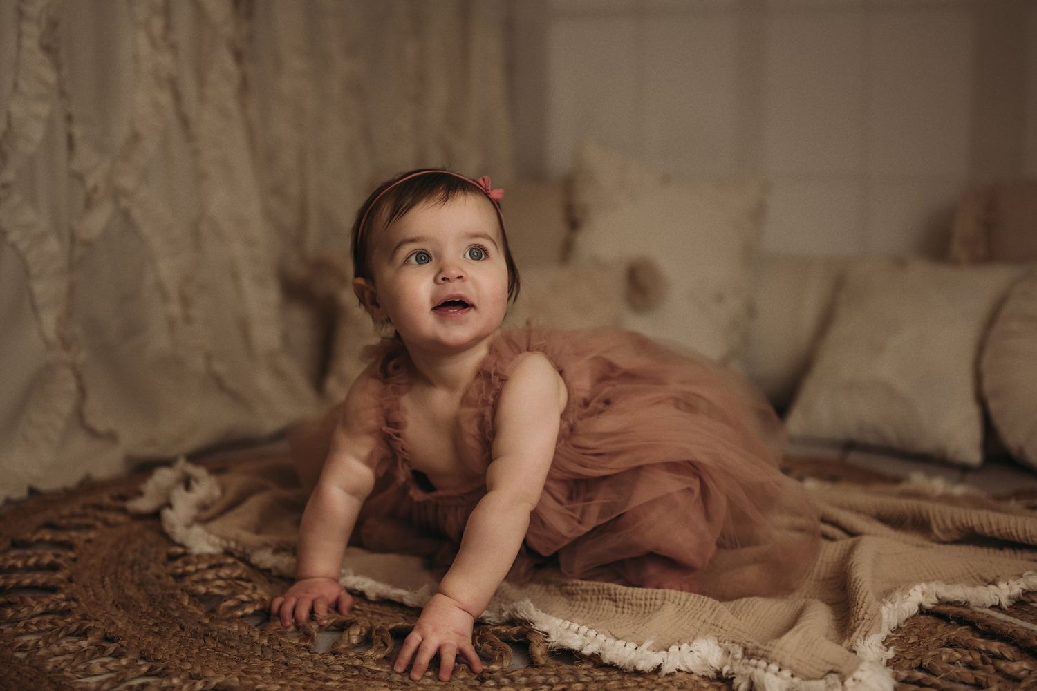 Toddler in dusty rose dress playing on textured rug with decorative pillows in background.
