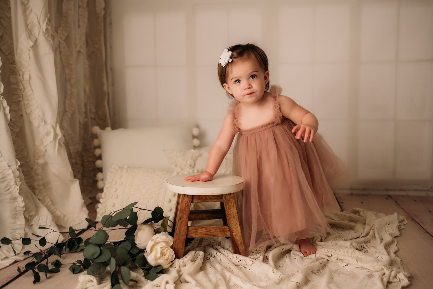 Baby in flowing mauve dress standing near wooden stool with white roses and greenery.