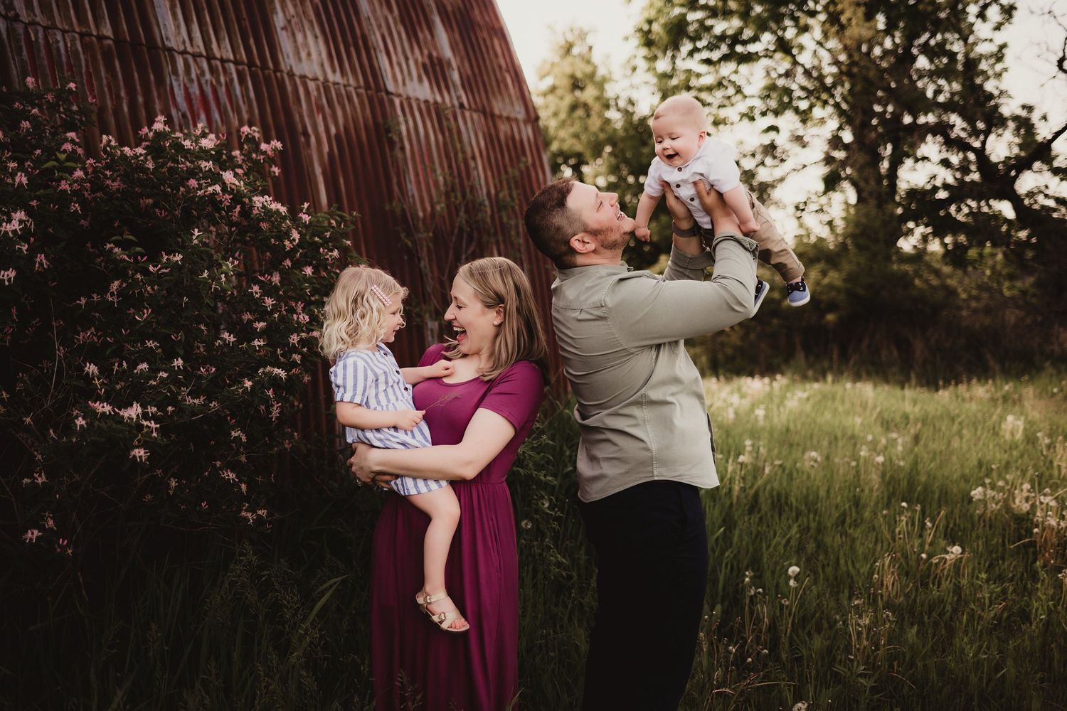 Family portrait in field shows heartwarming interaction near rustic barn structure.